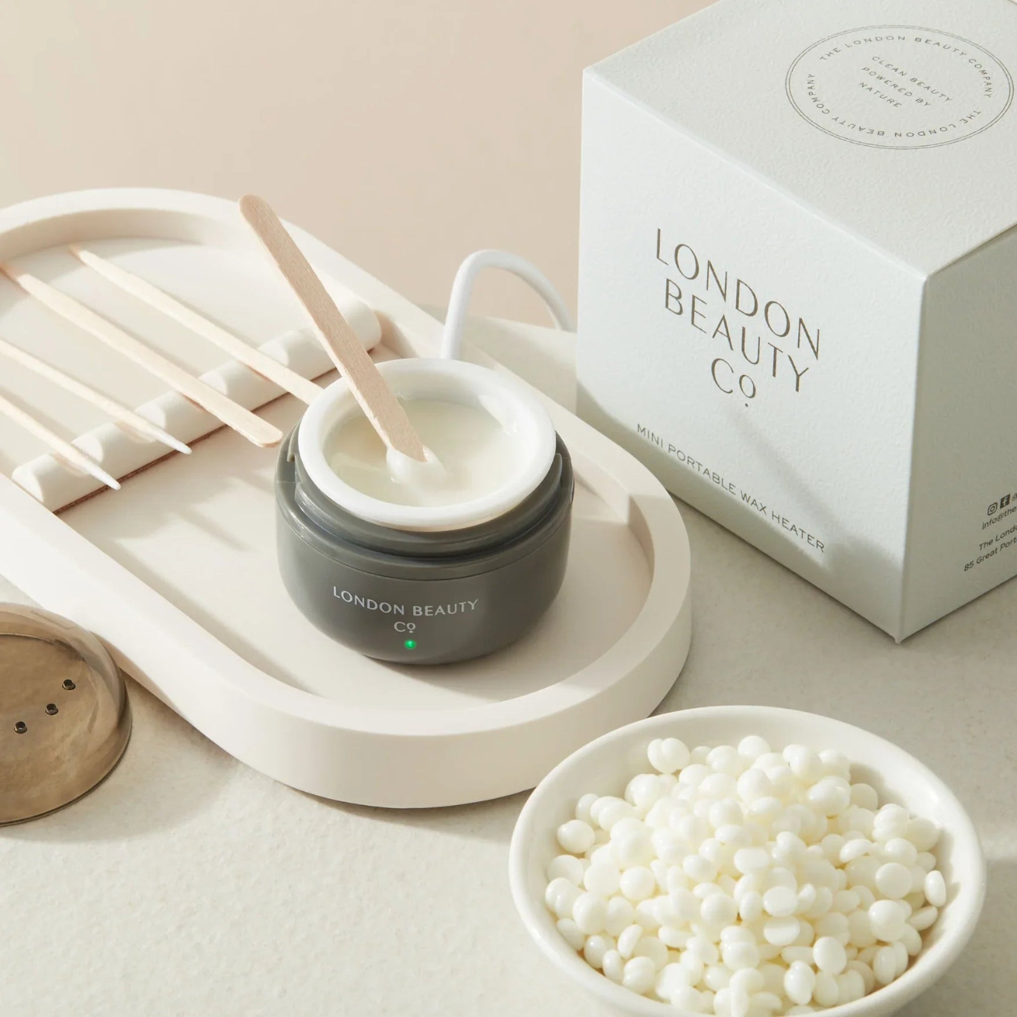 London Beauty Co Mini Portable Wax Heater lifestyle flatlay showing dark grey USB-powered mini wax heater with white silicone insert and wooden spatula resting across the top alongside its white London Beauty Co branded retail box a bowl of white wax pellets and wooden applicator sticks on a white oval tray from diagonal angle on warm cream background