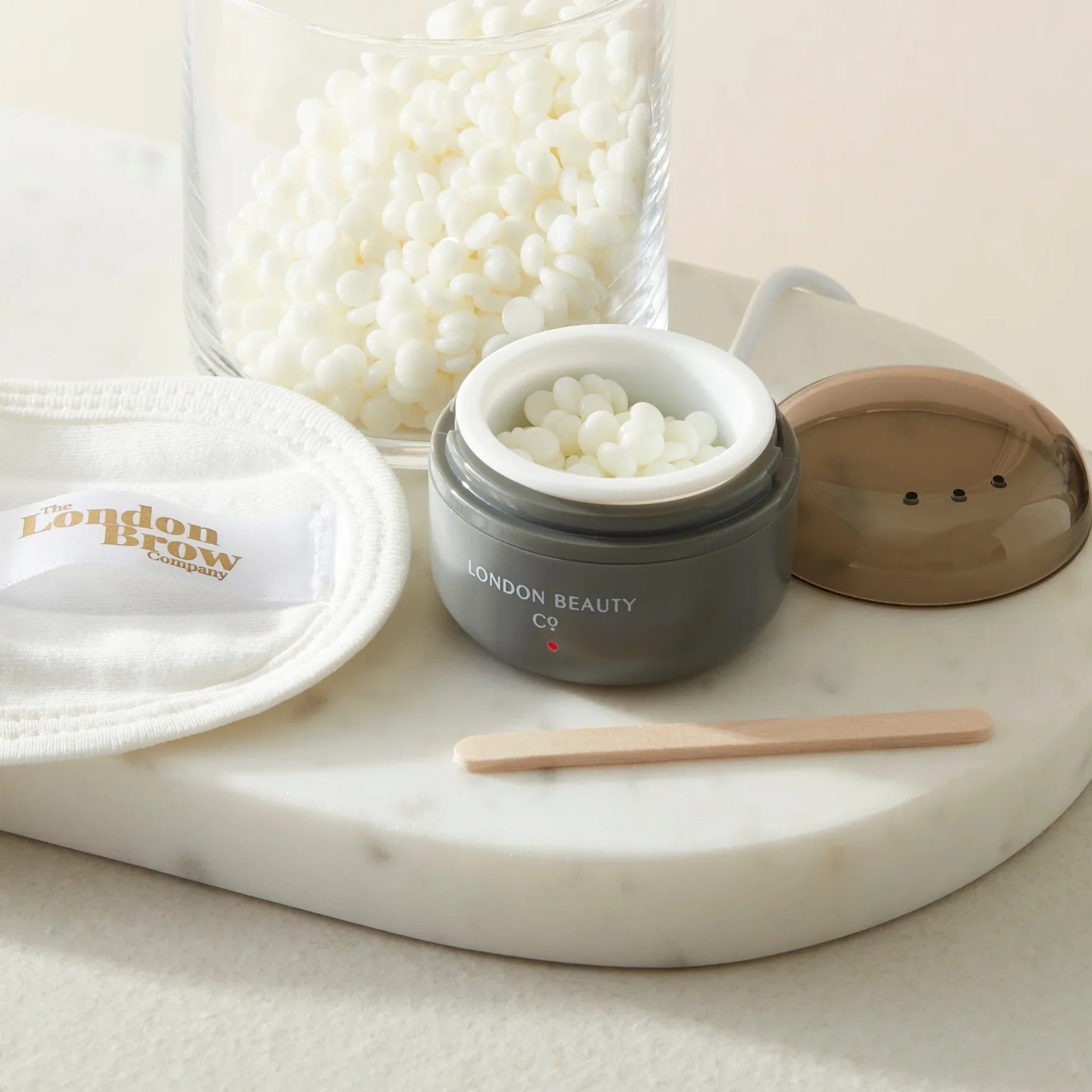 London Beauty Co Mini Portable Wax Heater lifestyle product shot showing dark grey USB-powered mini wax heater open with white wax pellets inside alongside its brown dome lid a glass jar of white wax pellets a wooden spatula and a London Brow Company branded reusable cotton pad on a white marble oval tray from diagonal angle on warm cream background