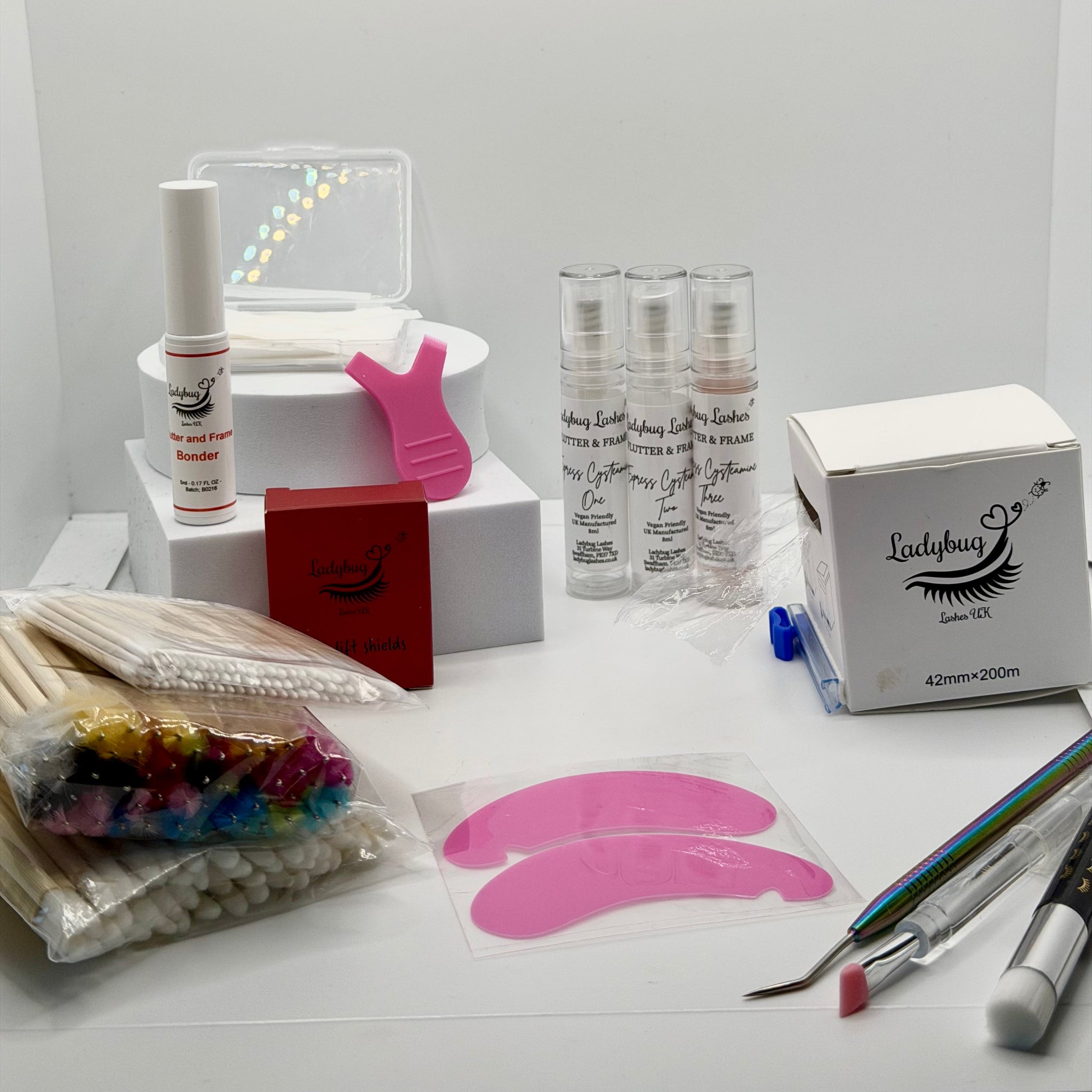 Ladybug Lashes Lash Lift Kit flatlay showing full kit contents including three Flutter and Frame Express Cysteamine treatment bottles, lash shampoo foam pump, coloured mascara wands, wooden spatulas, cotton buds, metal lash rod tool, white silicone lash shields, white medical tape roll and Ladybug Lashes branded cotton pad case on a white circular plinth on white background