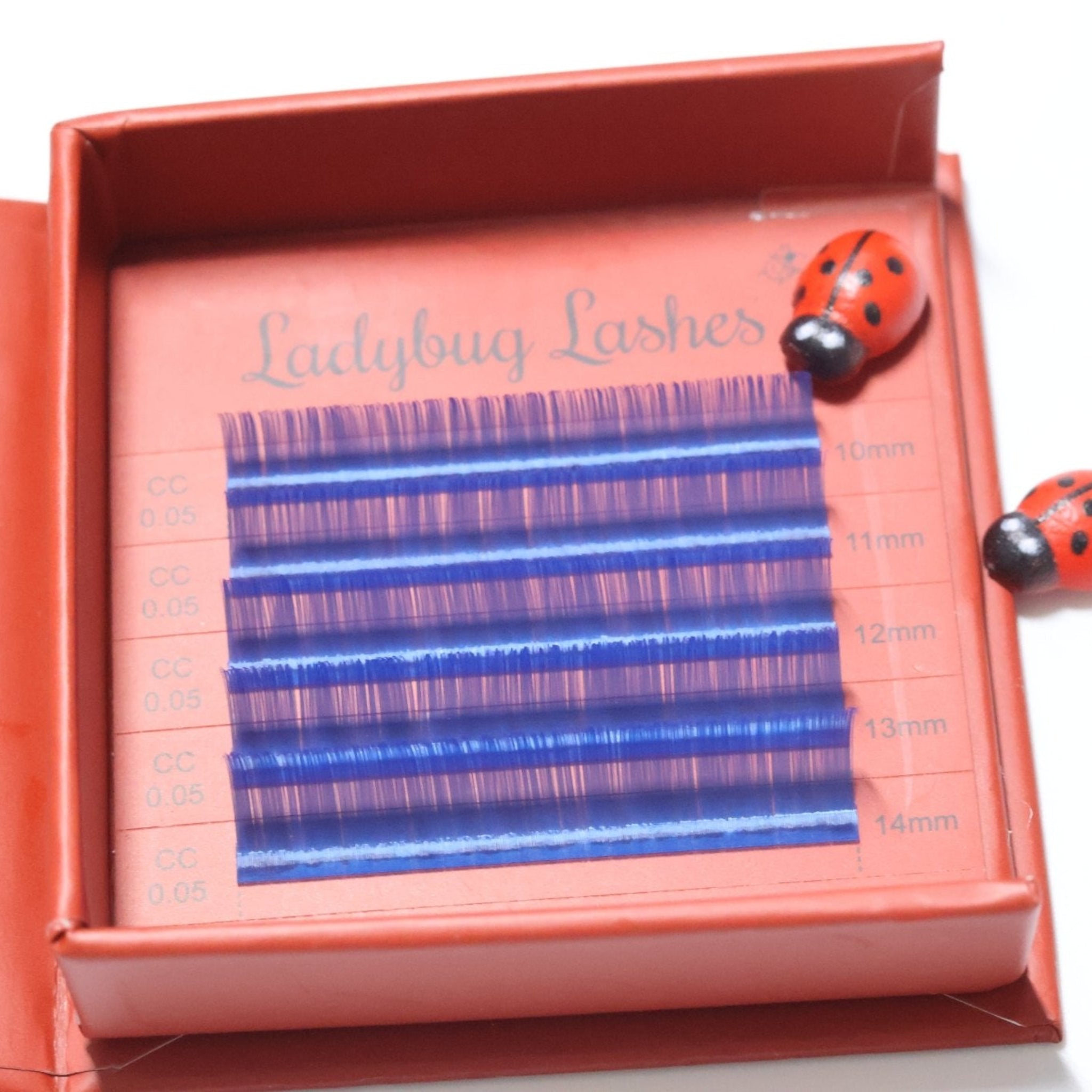 Ladybug Lashes Colour Volume Lashes 0.05mm open tray product shot showing multiple rows of blue and purple volume lash extensions in mixed lengths 10–14mm in a red Ladybug Lashes branded lash tray with two ceramic ladybug figurines from diagonal angle on white background