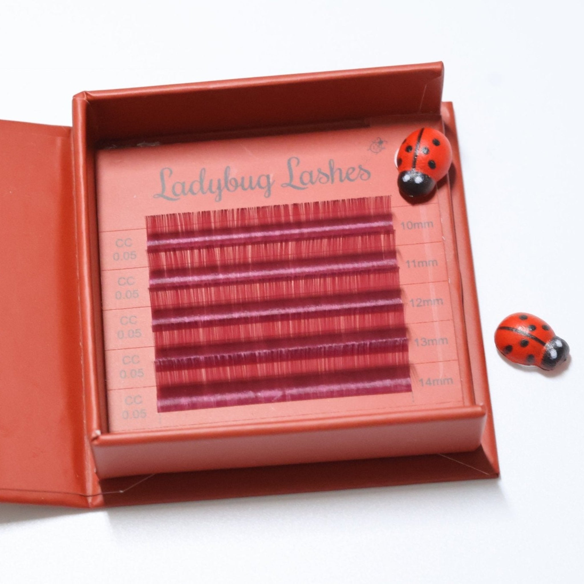 Ladybug Lashes Colour Volume Lashes 0.05mm open tray product shot showing multiple rows of red and pink volume lash extensions in mixed lengths 10–14mm in a red Ladybug Lashes branded lash tray with ceramic ladybug figurine from diagonal angle on white background