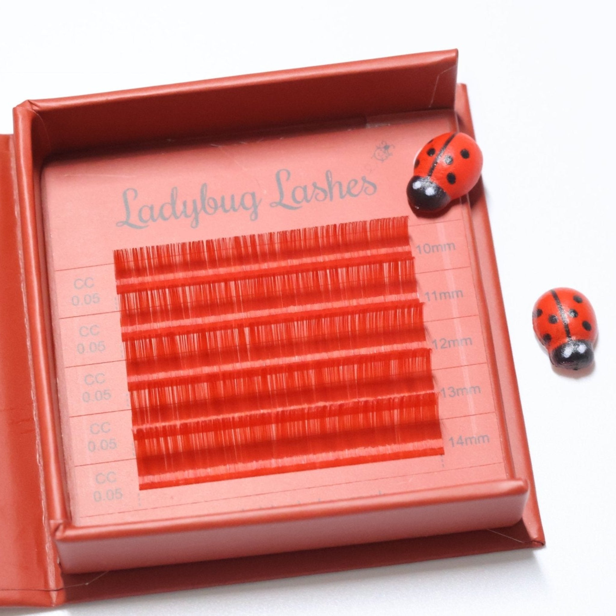 Ladybug Lashes Colour Volume Lashes 0.05mm open tray product shot showing multiple rows of red and orange volume lash extensions in mixed lengths 10–14mm in a red Ladybug Lashes branded lash tray with ceramic ladybug figurine from diagonal angle on white background