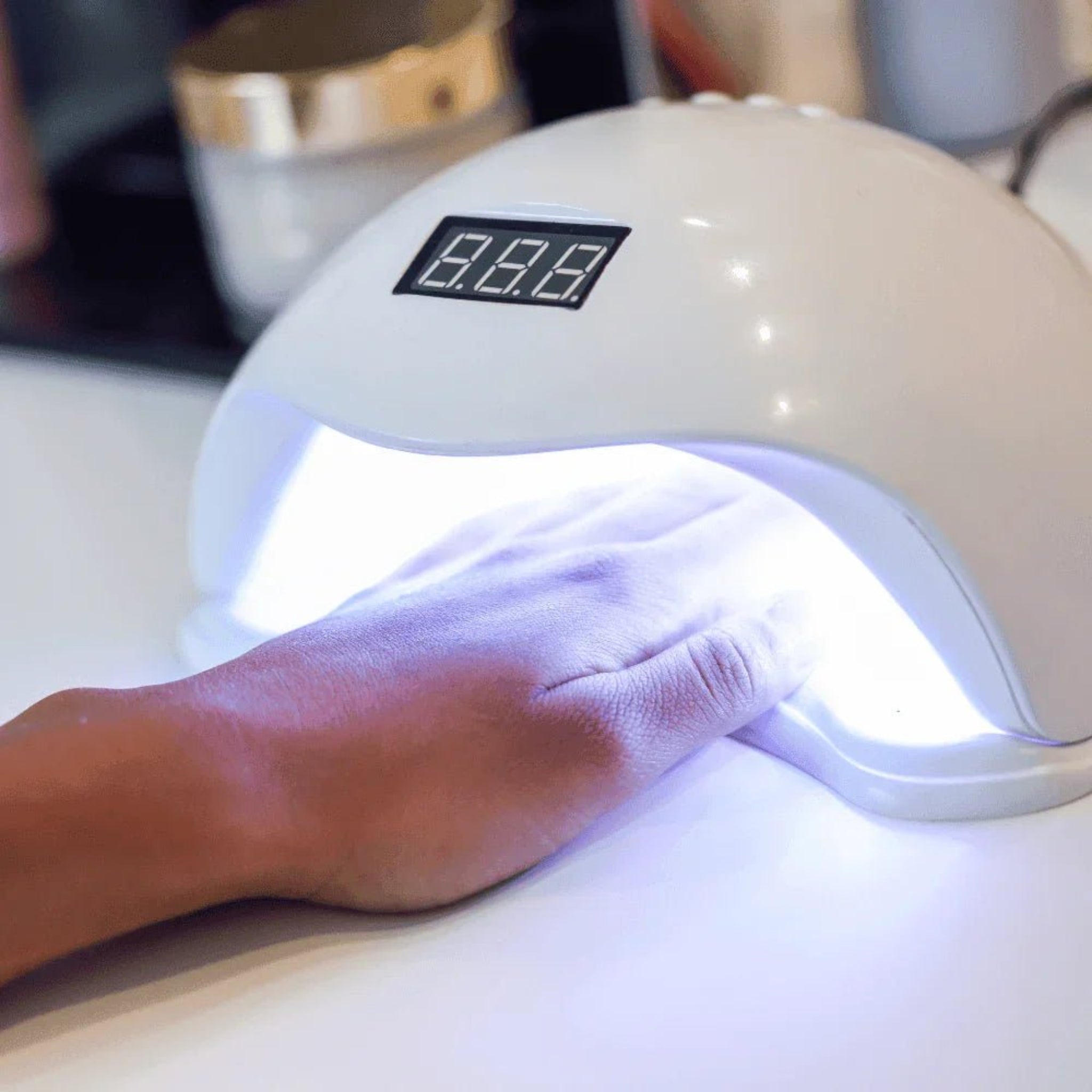 London Brow Company Gel UV Non-Chip Varnish Application Online Course in-use shot showing a medium-toned hand with gel-polished nails placed inside a white dome UV LED nail lamp with digital timer display illuminated from front angle on a white nail desk surface