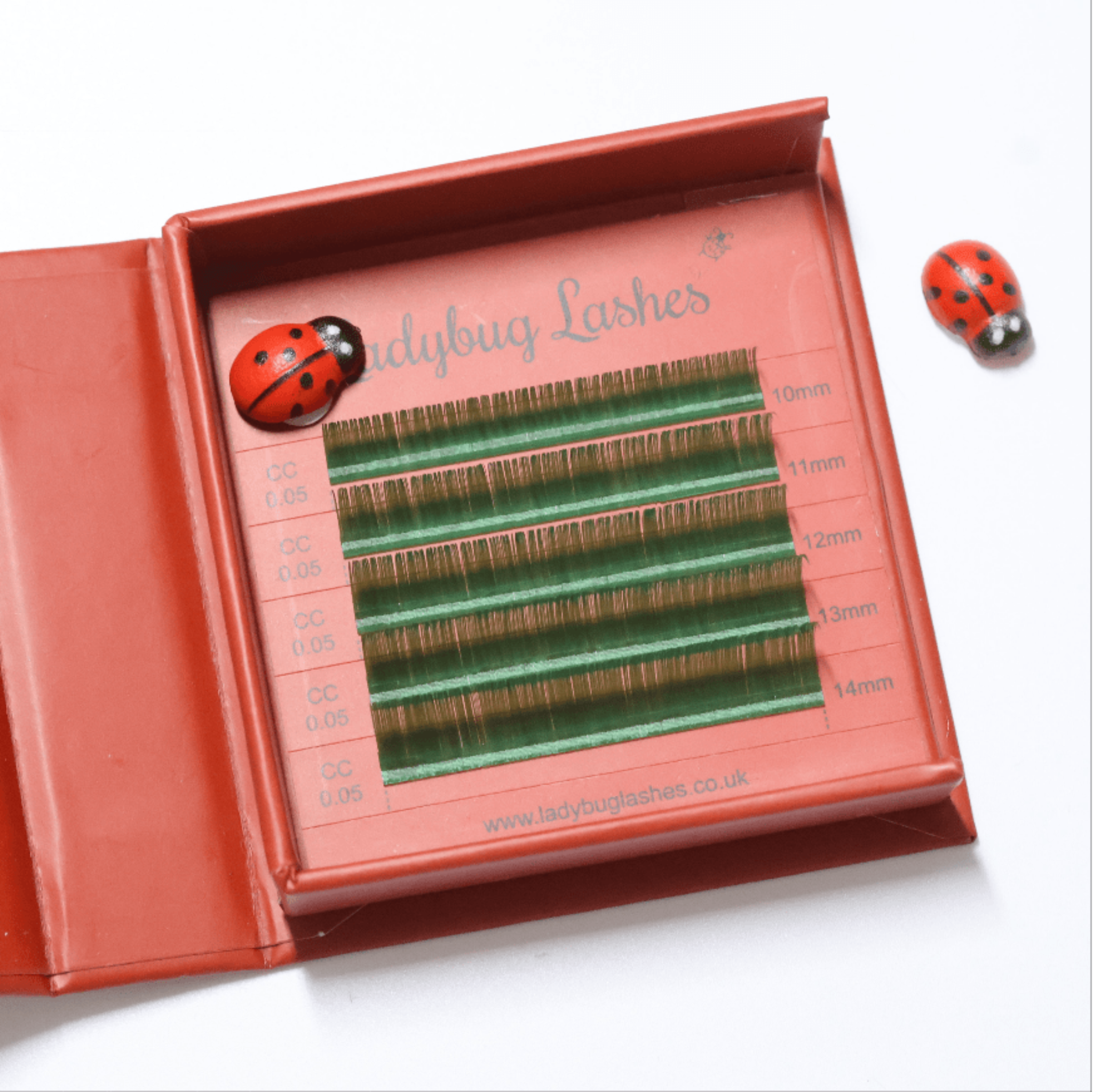 Ladybug Lashes Colour Volume Lashes 0.05mm open tray product shot showing multiple rows of green and gold volume lash extensions in mixed lengths 10–14mm in a red Ladybug Lashes branded lash tray with ceramic ladybug figurine from diagonal angle on white background