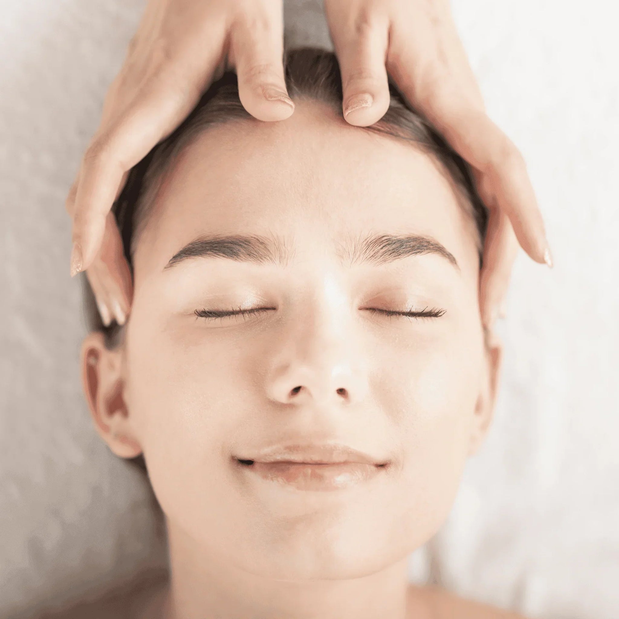 London Brow Company Indian Head Massage Online Course in-treatment overhead shot showing bare hands of a therapist applying gentle pressure to the forehead and temples of a relaxed fair-skinned female client with dark hair lying down with eyes closed and a soft smile from overhead angle on a cream spa background