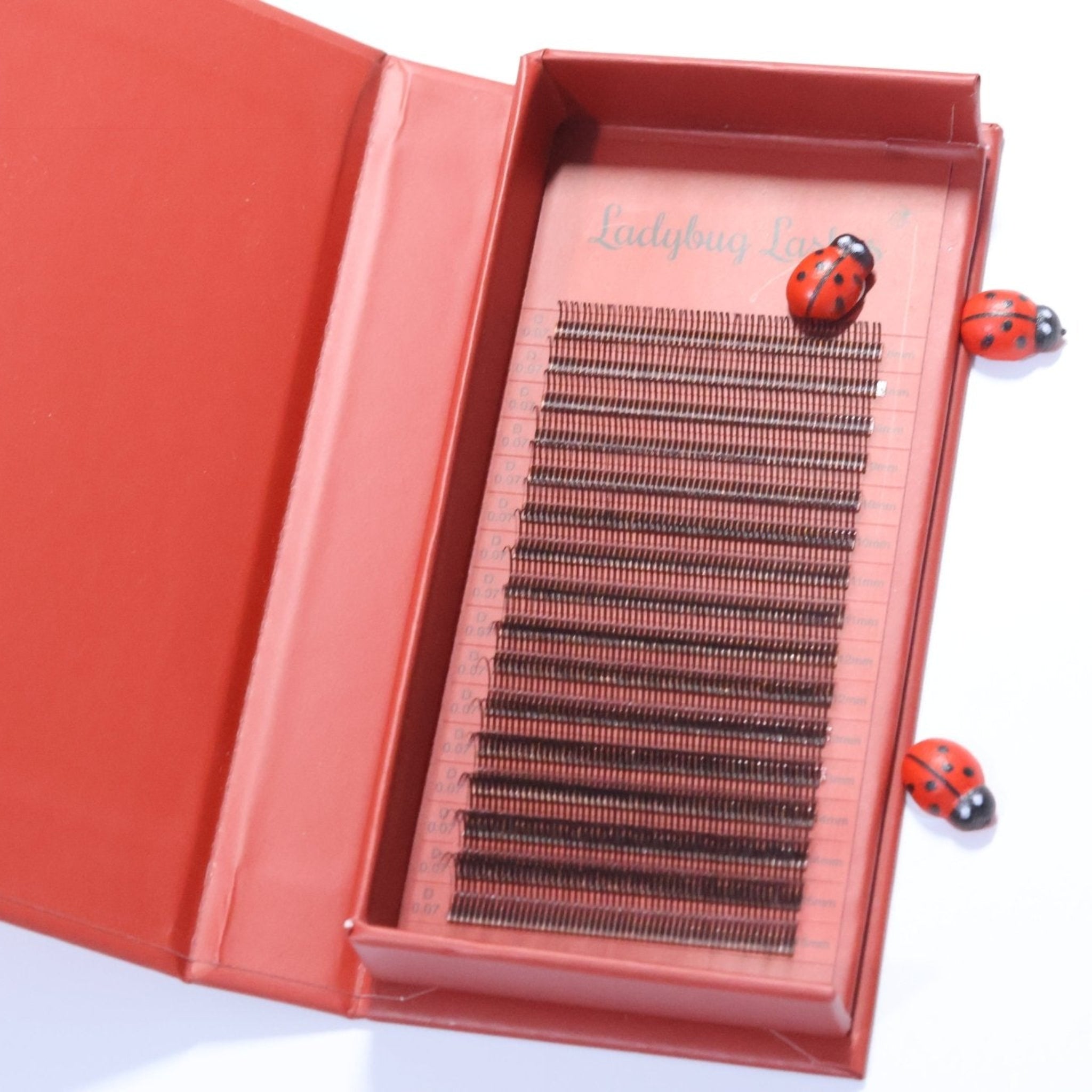Ladybug Lashes Brown Angel Lash 16 Row Mixed Tray overhead shot showing open red branded box with lid extended and 16 rows of warm brown angel lashes on a red tray insert, with Ladybug Lashes script branding and three decorative ladybird props visible