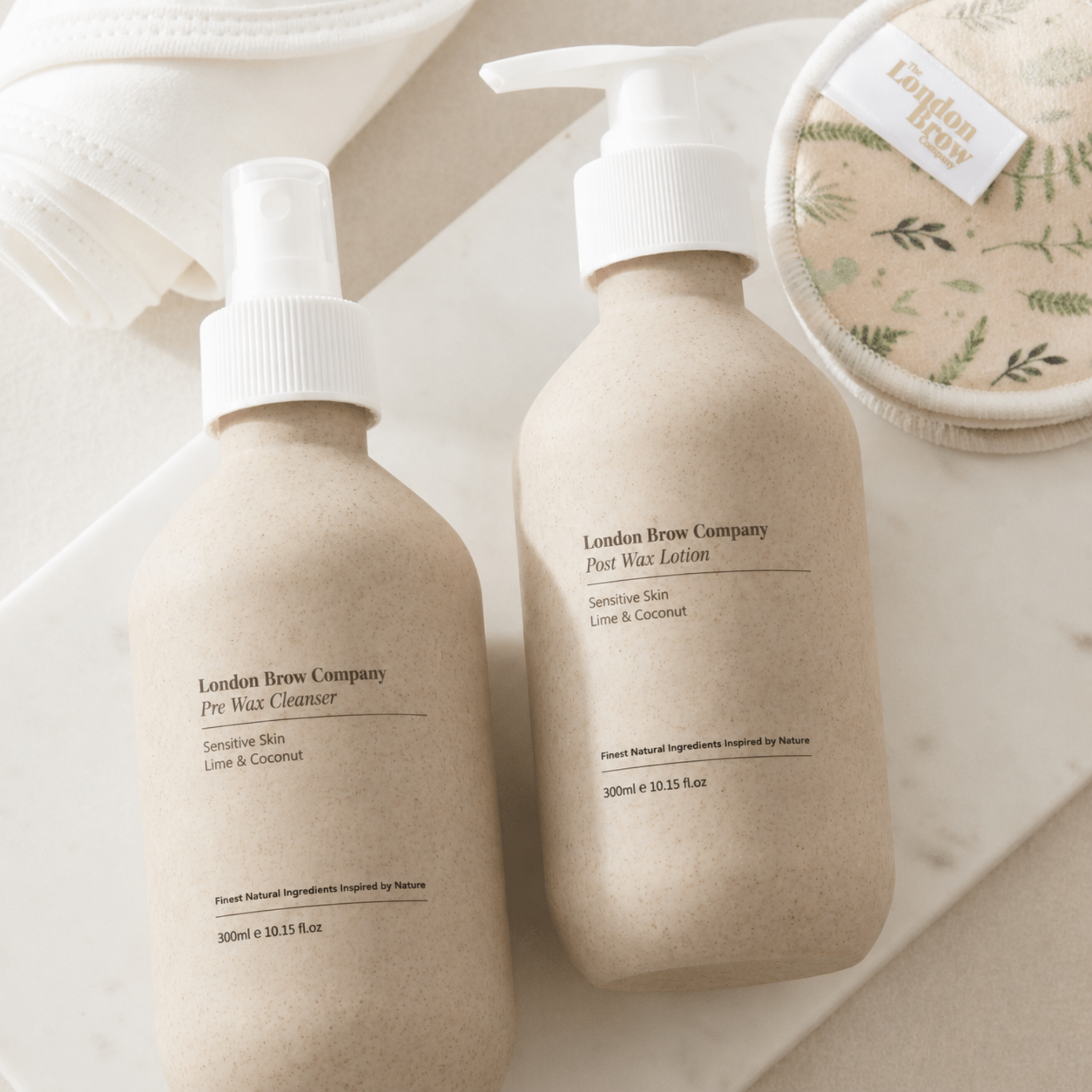The London Brow Company Pre-Wax Cleanser & Post-Wax Lotion Set flatlay product shot showing two 300ml stone-finish pump bottles labelled Pre Wax Cleanser and Post Wax Lotion Sensitive Skin Lime & Coconut alongside a London Brow Company branded reusable cotton pad from diagonal overhead angle on white marble surface
