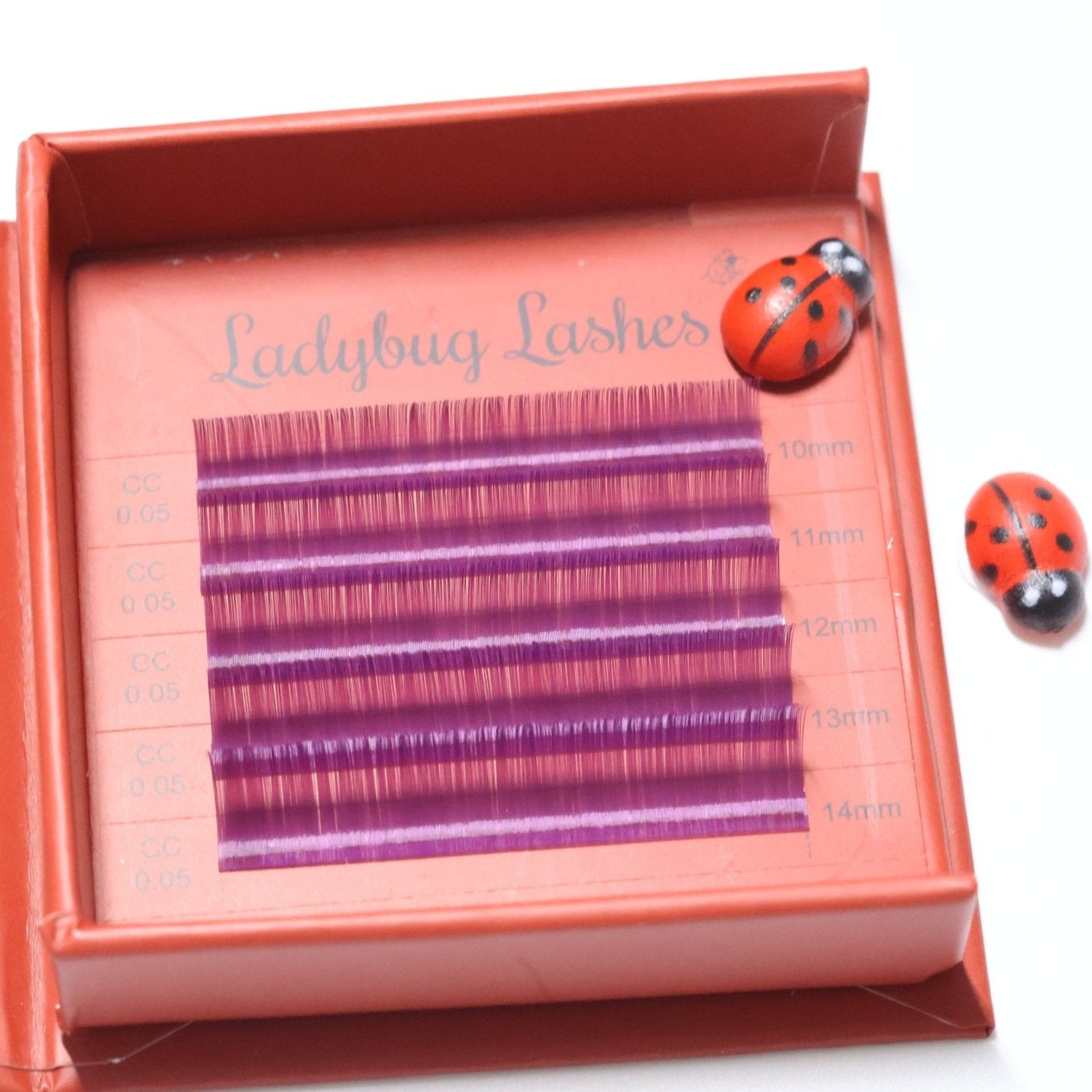 Ladybug Lashes Colour Volume Lashes 0.05mm open tray product shot showing multiple rows of purple and pink volume lash extensions in mixed lengths 10–14mm in a red Ladybug Lashes branded lash tray with ceramic ladybug figurine from diagonal angle on white background