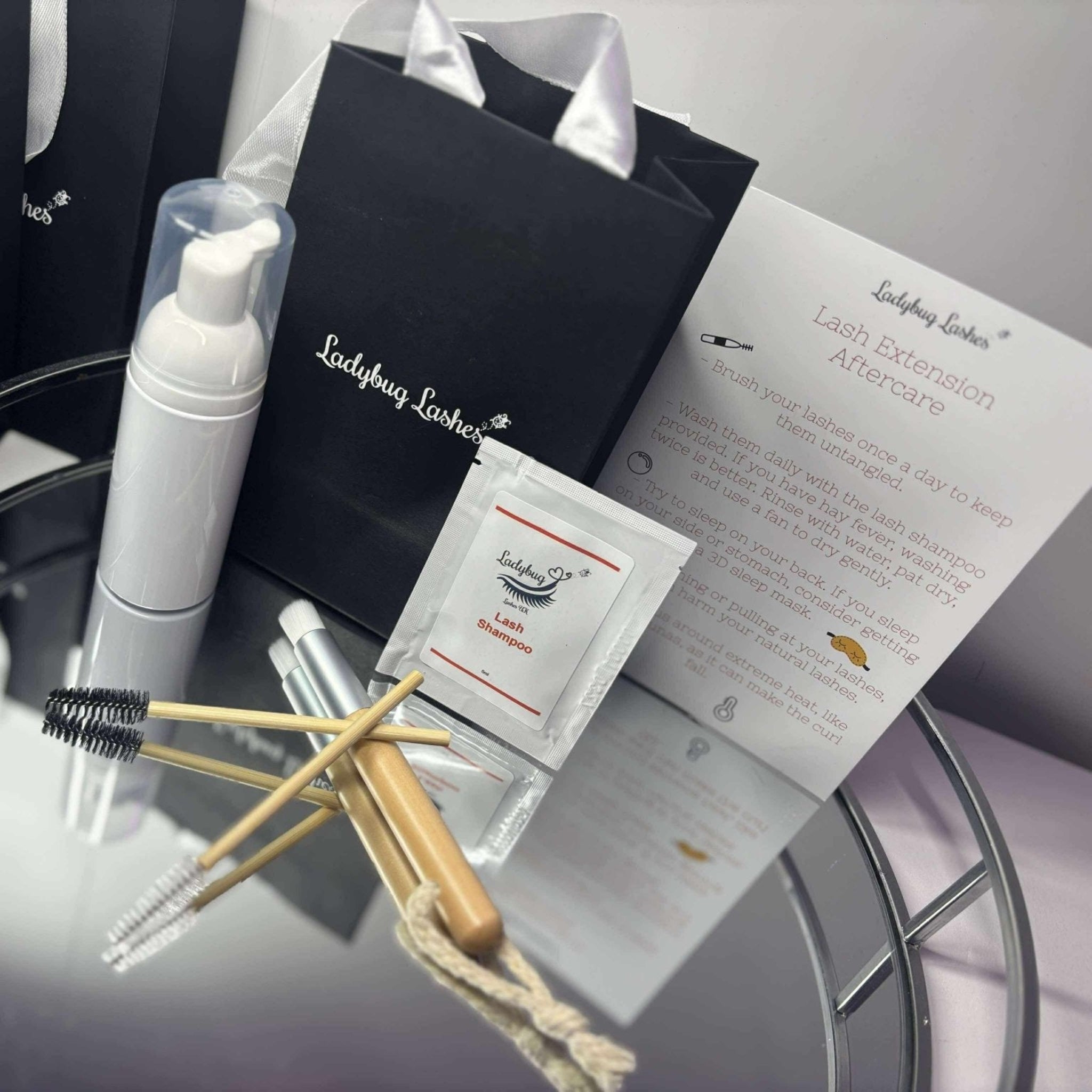 Overhead angle of Ladybug Lashes Lash Shampoo Sachet Kit contents arranged on a glass surface in front of multiple black Ladybug Lashes gift bags with white tissue paper, including a white foam pump bottle, a lash shampoo sachet, a bamboo spoolie, a bamboo cleansing brush, a mini applicator, and a printed Lash Extension Aftercare card