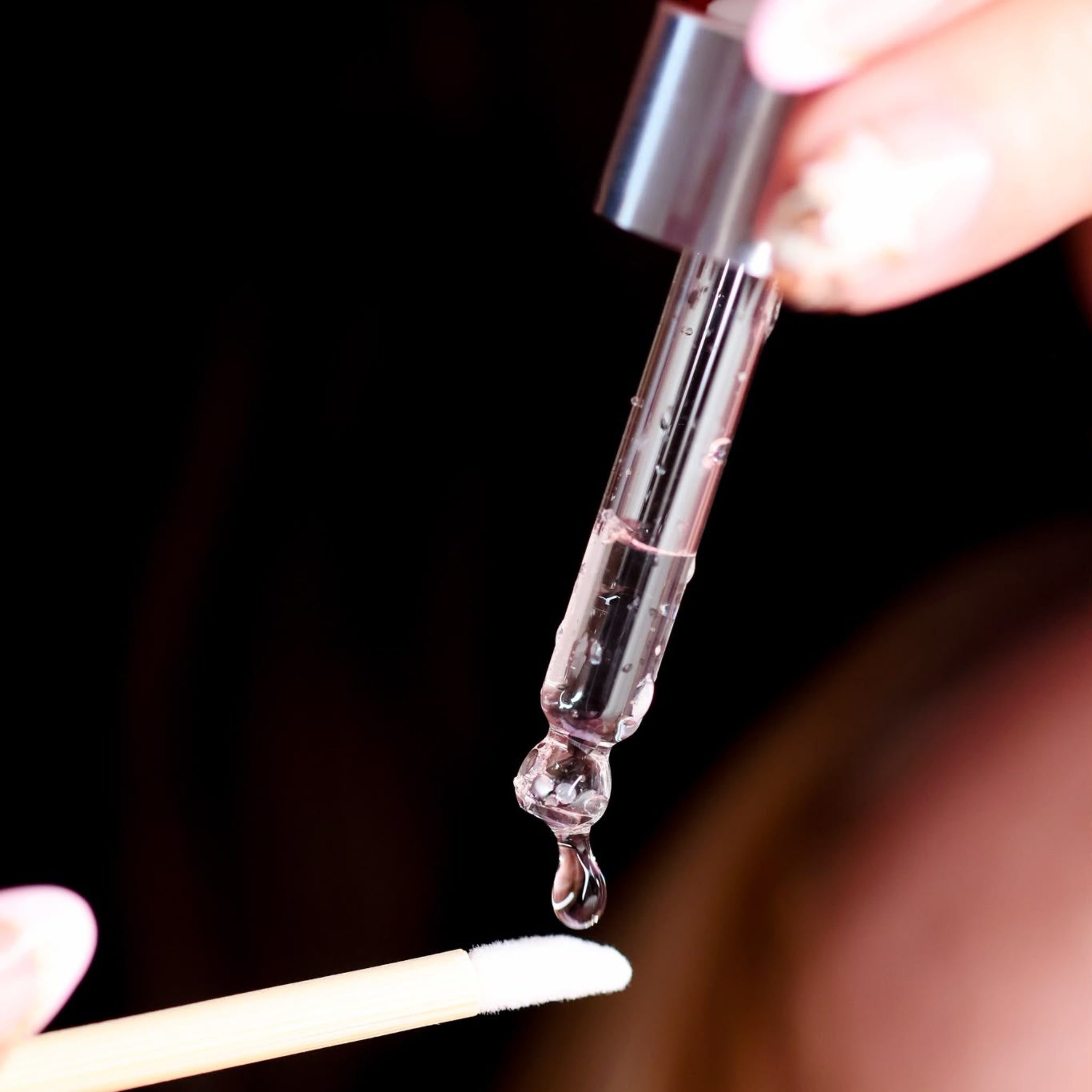 A close-up of a clear glass dropper with a silver cap dispensing a drop of transparent liquid onto a small beige applicator stick, held by hands against a dark background.