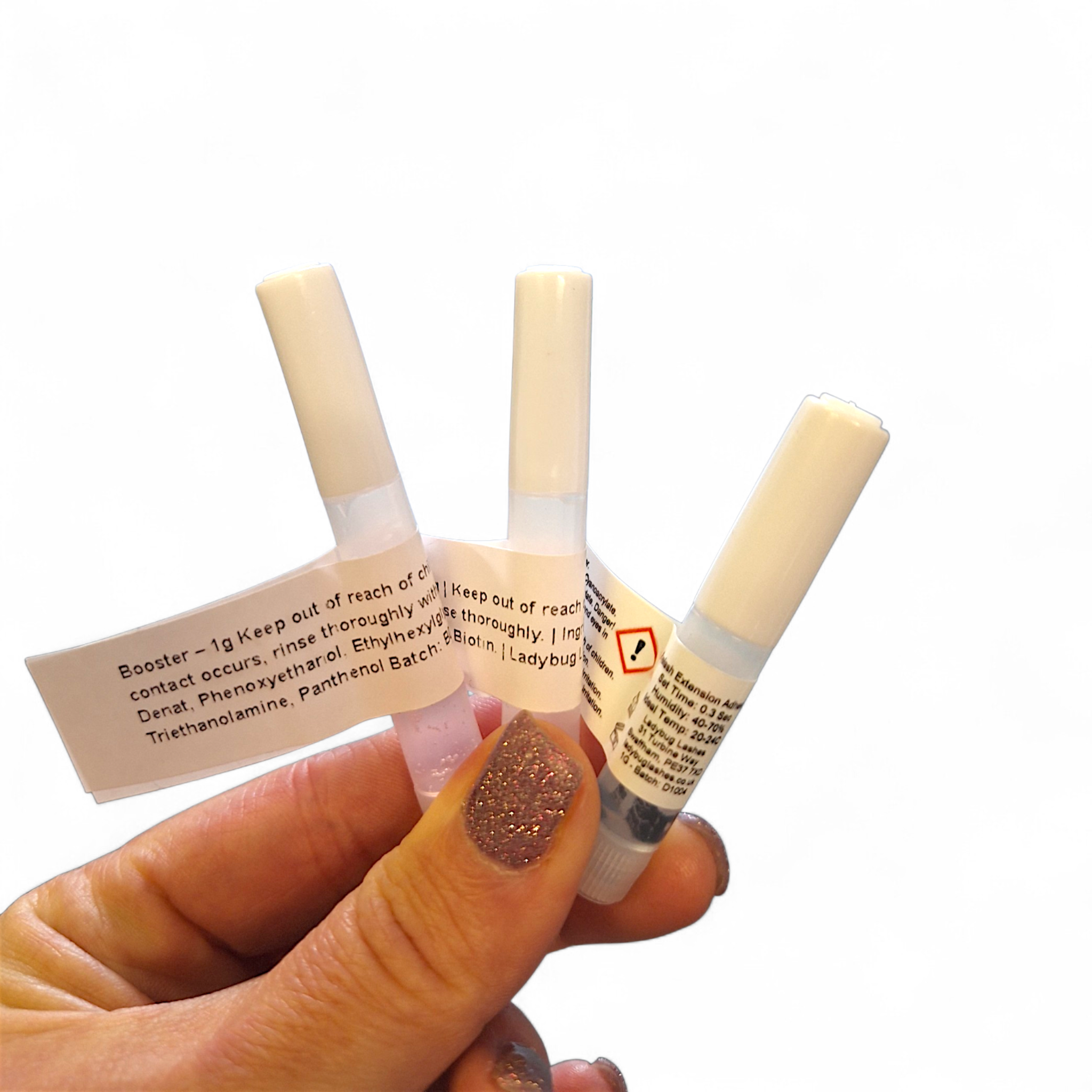 Hand with rose gold glitter nail polish holding three Ladybug Lashes 1g sample bottles fanned out against a white background — a Booster sample with white label showing ingredients including Denat, Phenoxyethanol, Ethylhexylglycerine, Triethanolamine and Panthenol, a SuperBonder sample with white label, and a lash extension adhesive sample bottle with hazard warning label and Ladybug Lashes branding visible