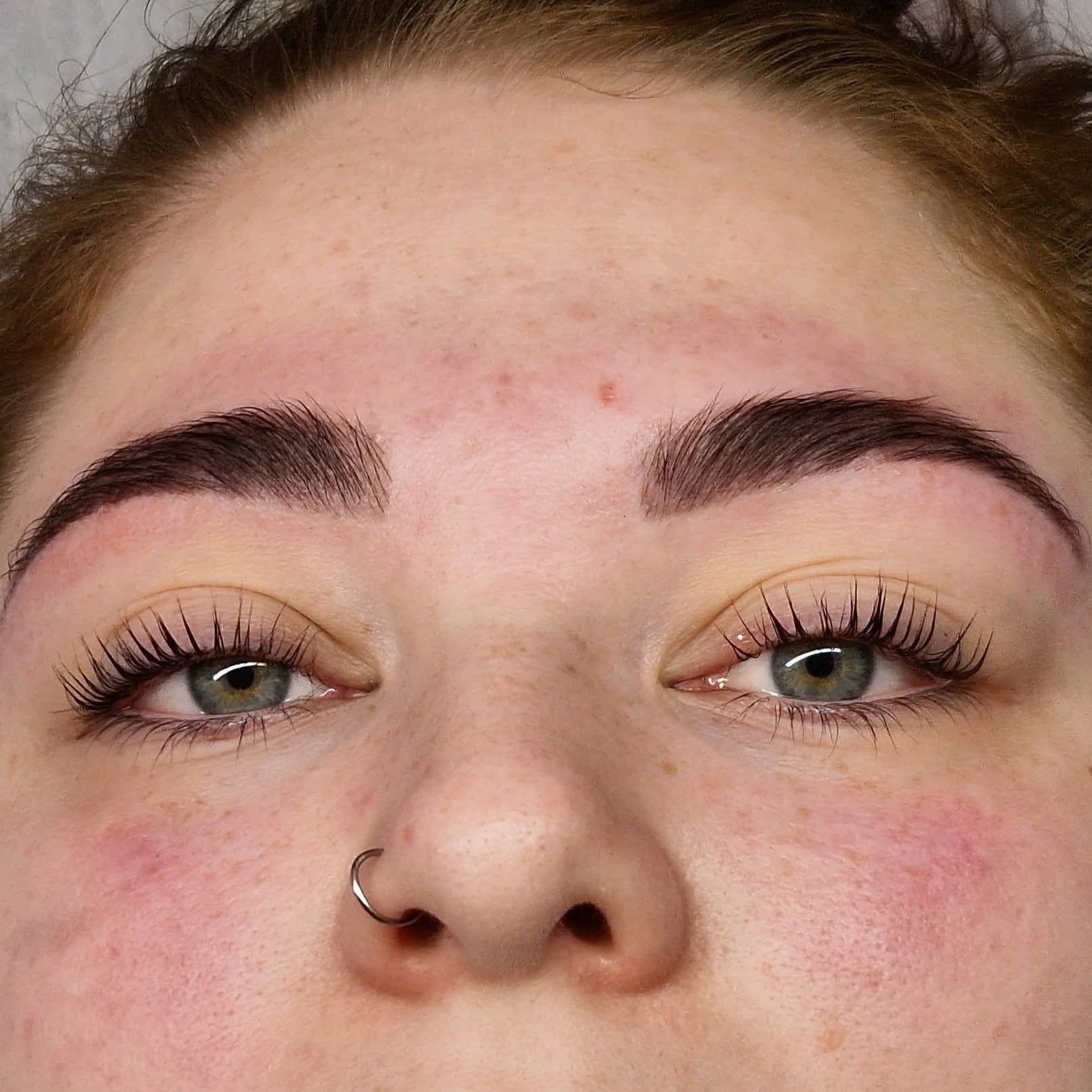 Ladybug Lashes Airless Pump Express Cysteamine Brow Lamination Kit treatment result shot showing model lying down with lifted lashes and freshly laminated full dark brows from front overhead angle with grey-green eyes and nose ring visible