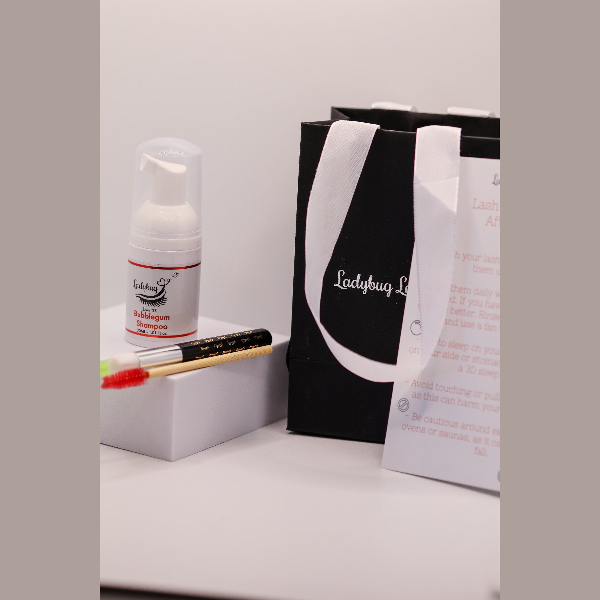 Ladybug Lashes Lash Shampoo Kit contents showing a Ladybug Lashes Bubblegum Shampoo 30ml foam pump bottle on a white plinth, a bamboo-handled lash cleansing brush with red spoolie, a black Ladybug Lashes gift bag with white ribbon handles and a printed Lash Extension Aftercare card on a white surface