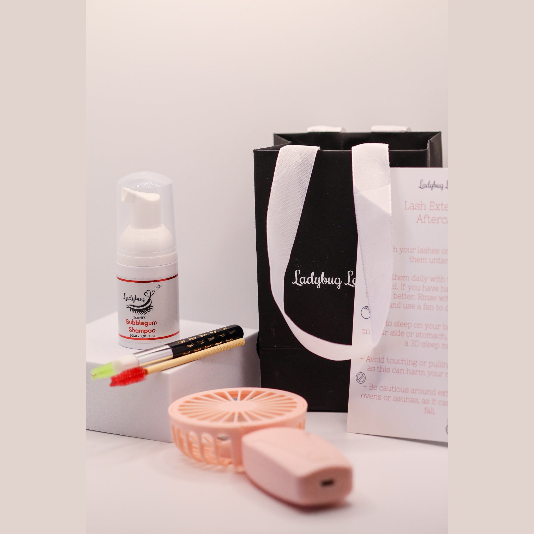 Ladybug Lashes Lash Shampoo Kit with Fan contents showing a Ladybug Lashes Bubblegum Shampoo 30ml foam pump bottle on a white plinth, a bamboo-handled lash cleansing brush with red spoolie, a pink portable USB lash drying fan, a black Ladybug Lashes gift bag with white ribbon handles and a printed Lash Extension Aftercare card on a white surface
