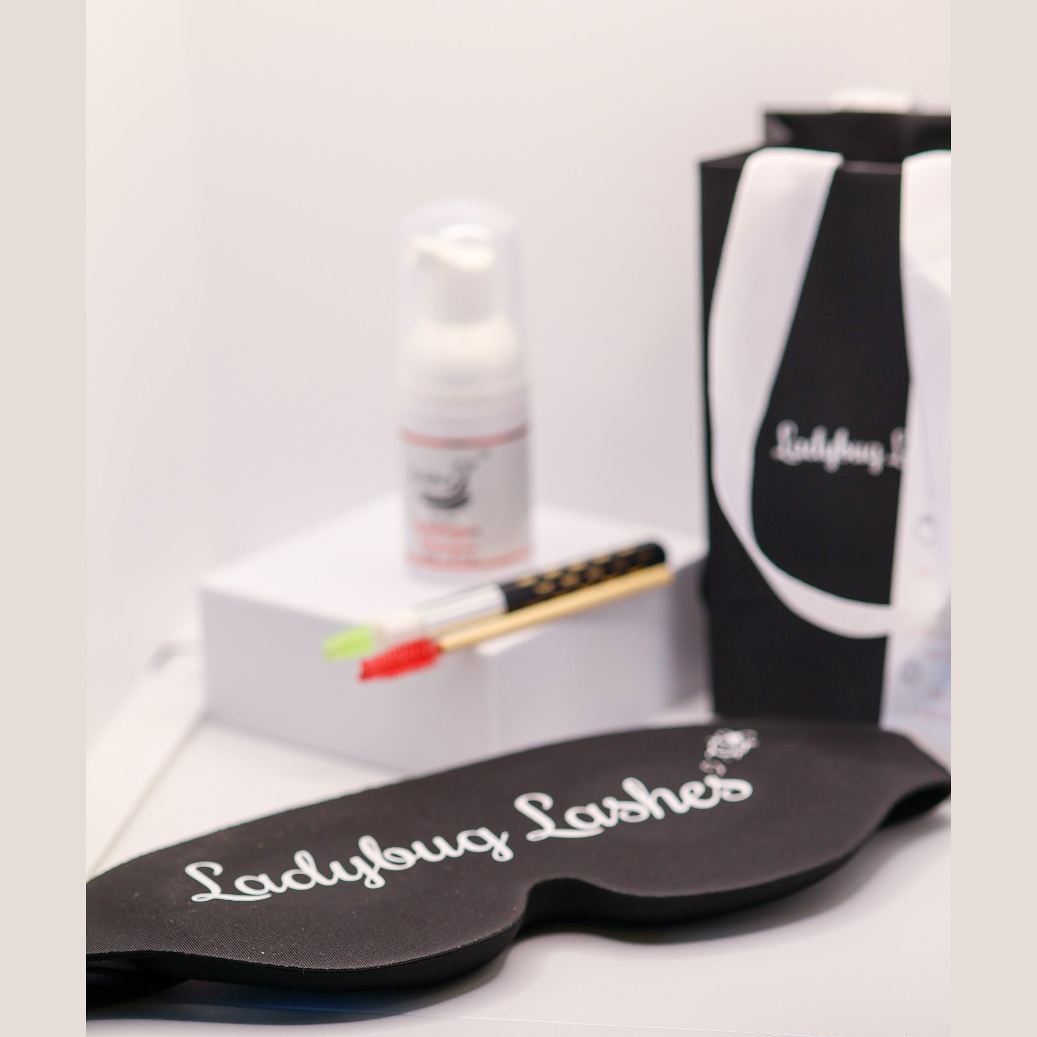 Ladybug Lashes Lash Shampoo Kit with Eye Mask contents showing a black Ladybug Lashes branded sleep mask in the foreground, a Ladybug Lashes Bubblegum Shampoo foam pump bottle and bamboo-handled lash cleansing brush with red spoolie on a white plinth, and a black Ladybug Lashes gift bag with white ribbon handles on a white surface