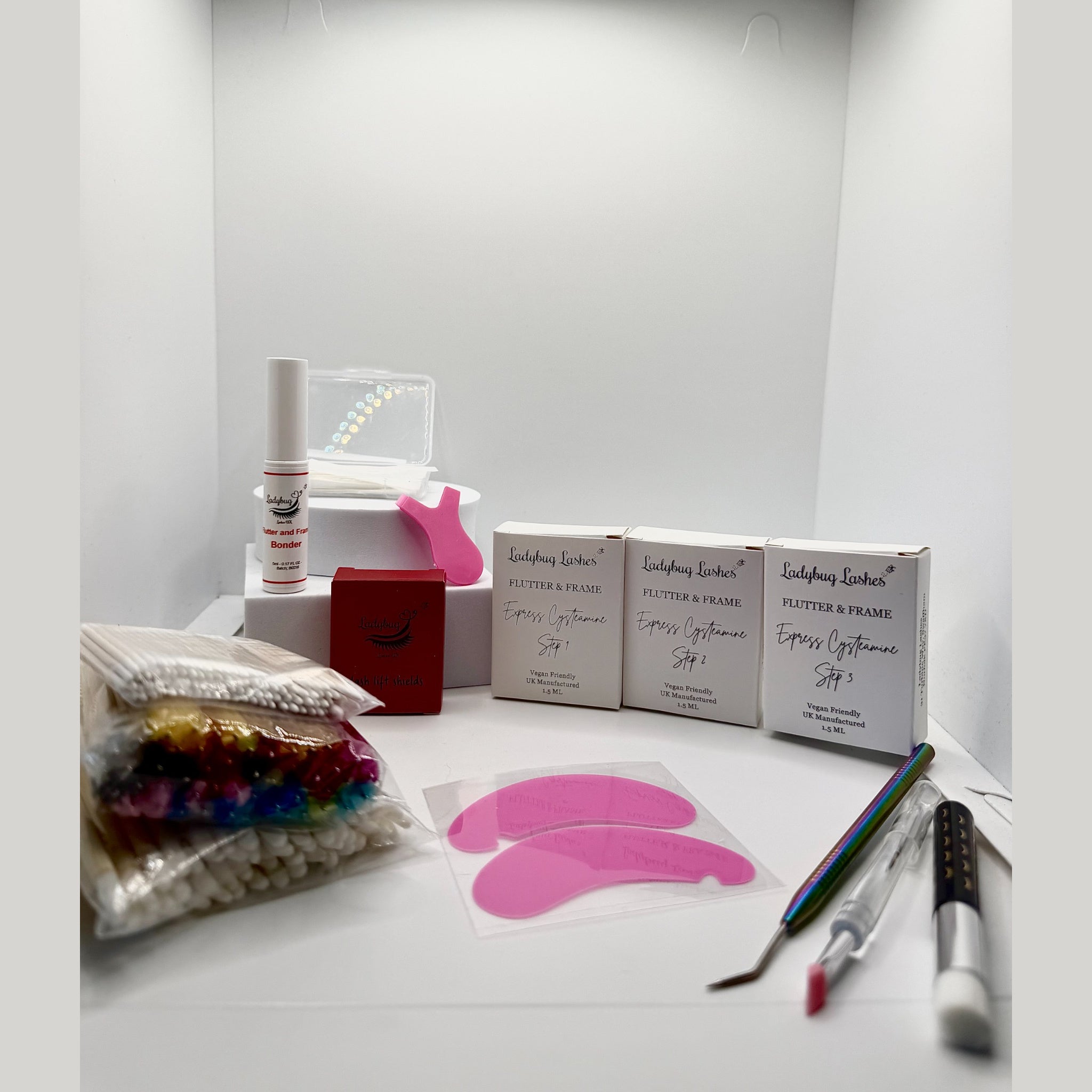 Ladybug Lashes Express Cysteamine Lash Lift Sachet Kit flatlay showing Flutter and Frame Step 1, 2 and 3 sachet boxes, Bonder bottle, pink lash lift tool, silicone under eye pads, rainbow mascara wands, microfibre brushes, tweezers and disposable applicators on white background