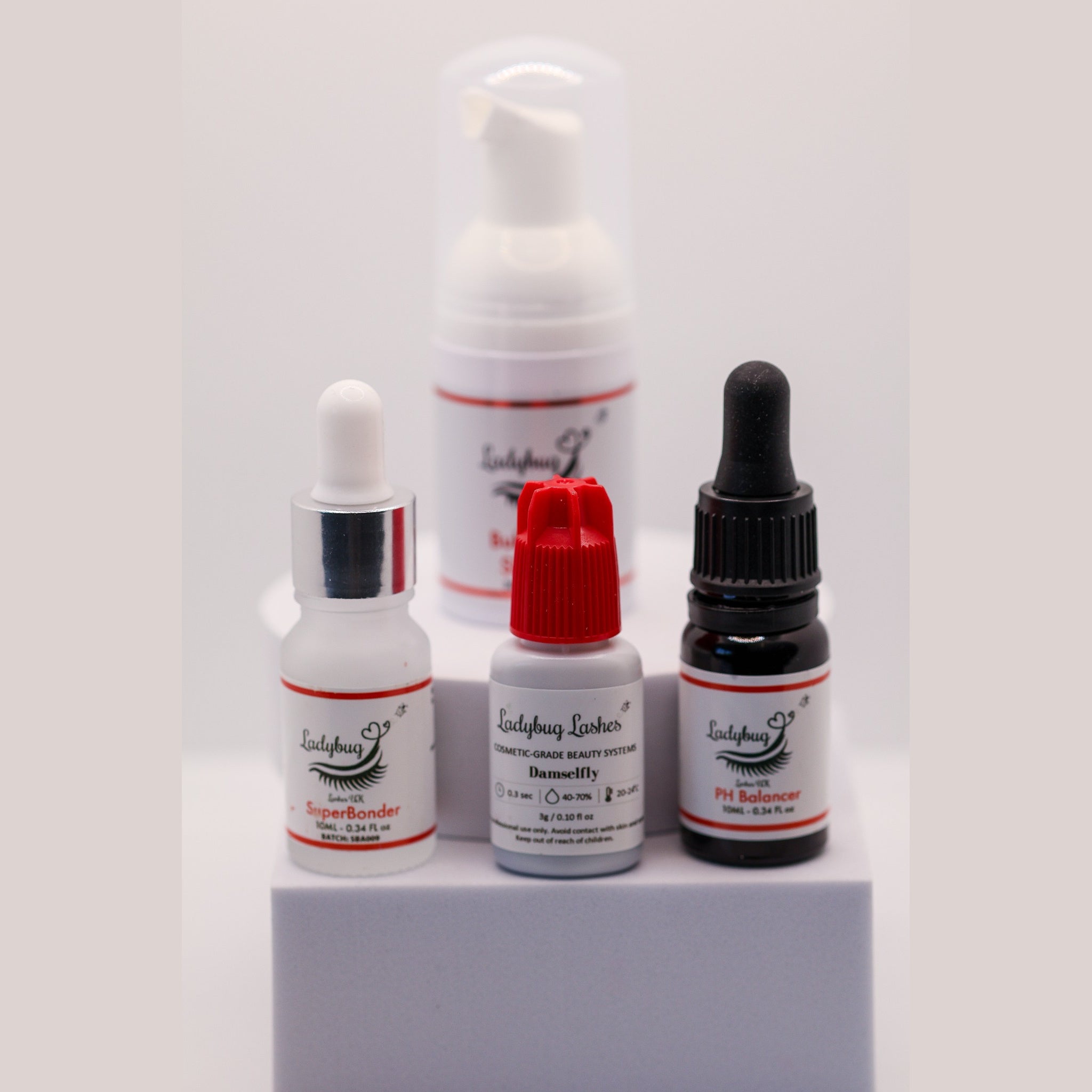 Ladybug Lashes Retention Trio showing four products on a white background: Bubblegum Shampoo 30ml foam pump bottle, SuperBonder 10ml clear glass dropper, PH Balancer 10ml dark amber dropper, and Wildflower UK adhesive 5g bottle with red cap