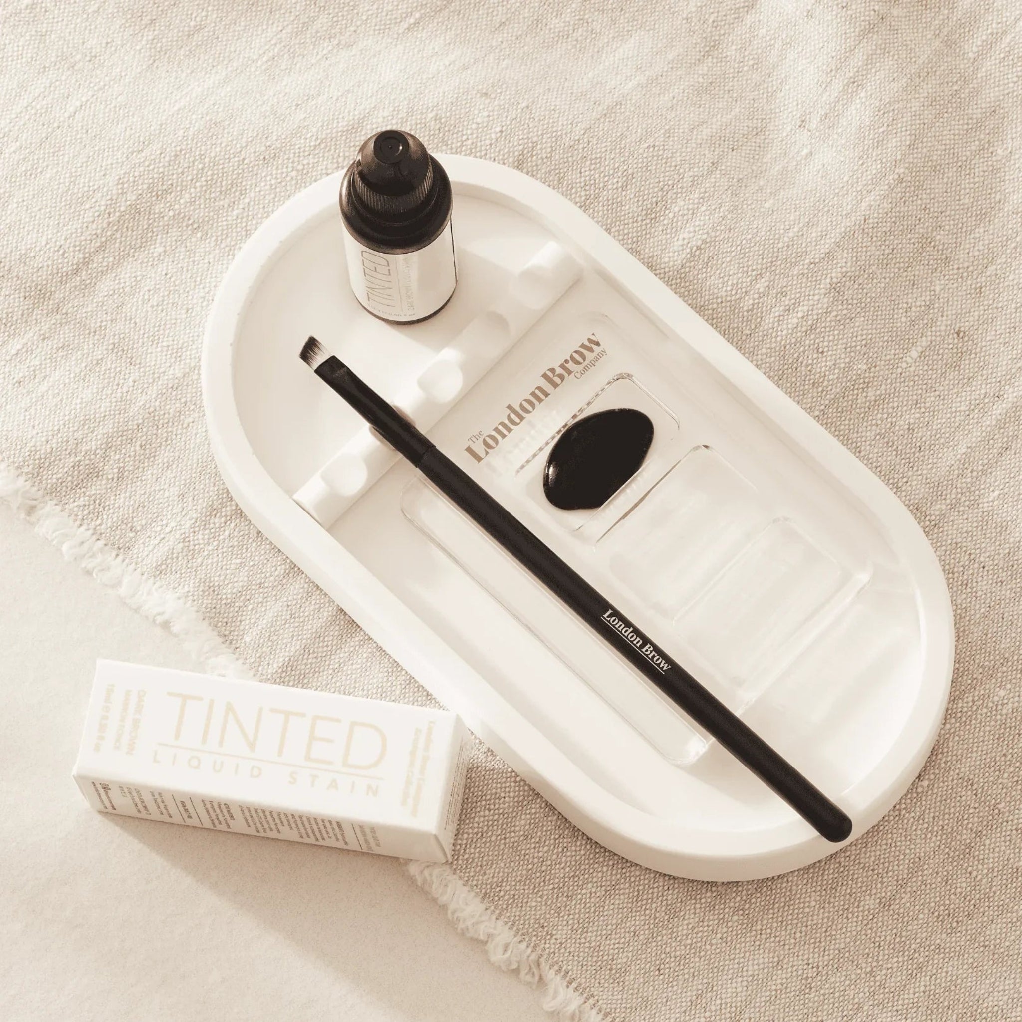 London Brow Company Kensington Collection Tinted Liquid Stain in-use lifestyle flatlay showing a dark amber dropper bottle a London Brow Company flat angled brow brush and a white London Brow Company ceramic mixing tray with a black mixed stain swatch on a cream linen surface from diagonal overhead angle