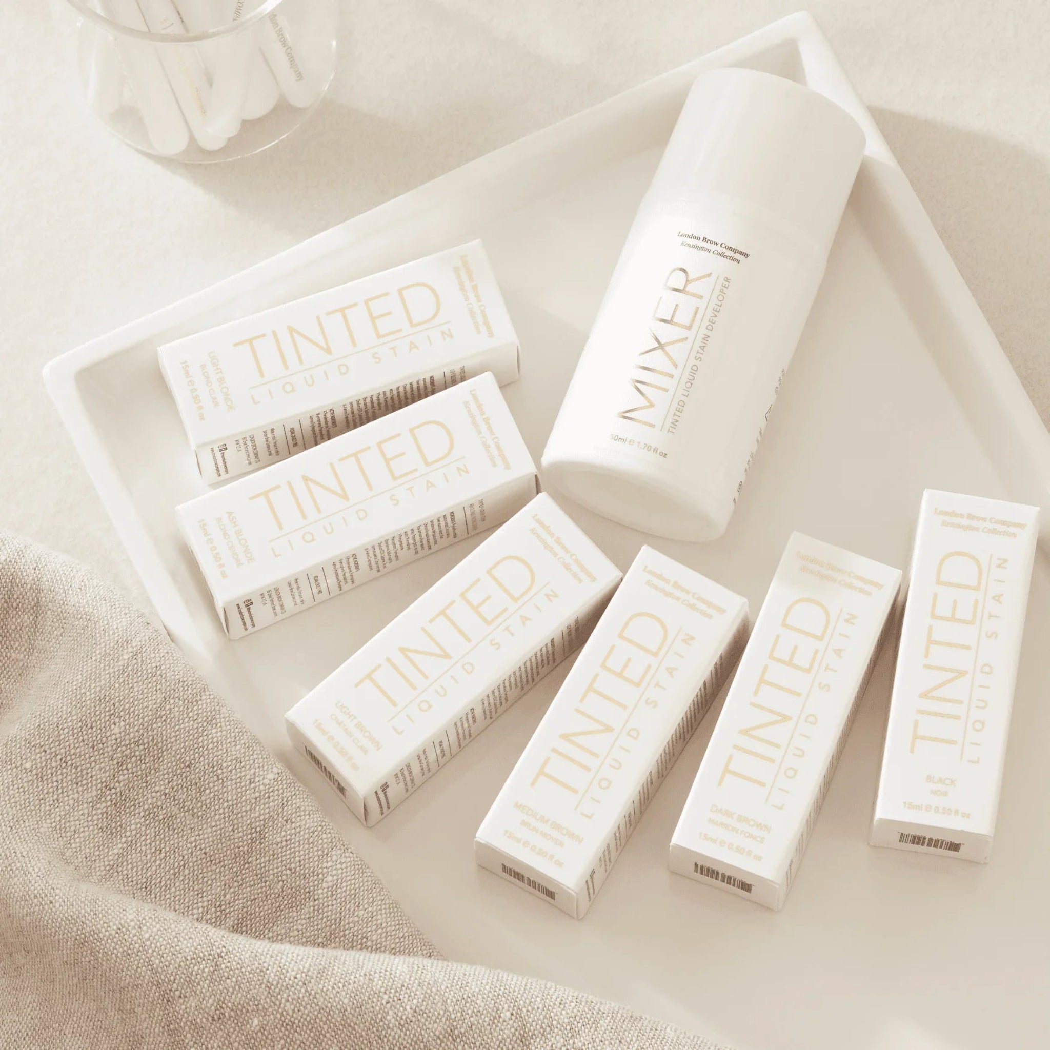 London Brow Company Kensington Collection Tinted Liquid Brow Stain 15ml full shade range flatlay showing seven white retail boxes labelled Tinted Liquid Stain in gold alongside a white London Brow Company Mixer bottle arranged on a white ceramic tray on a warm cream linen surface from diagonal overhead angle