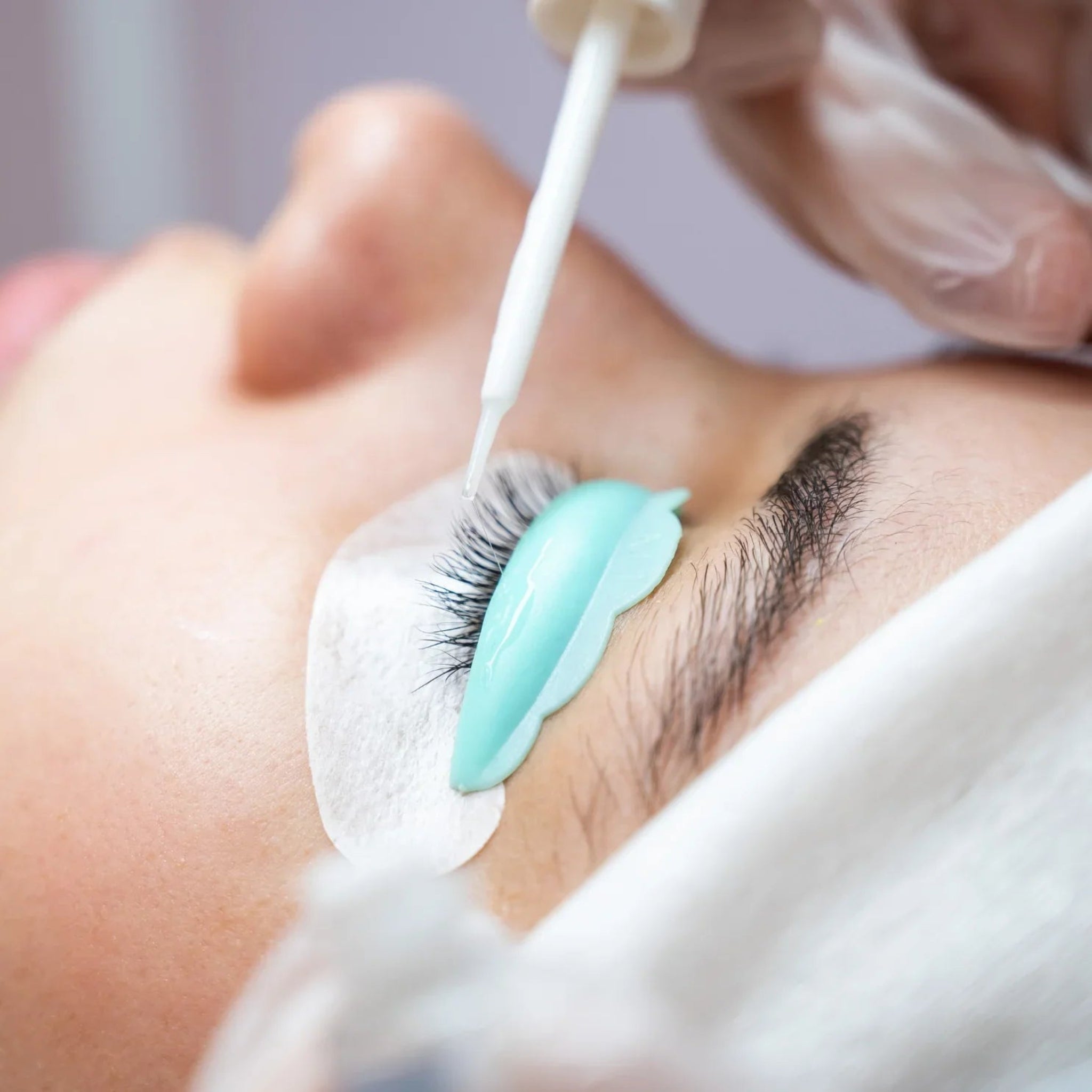 London Brow Company Speed-Lift Lash Lift and Tinting Course in-treatment extreme close-up showing gloved therapist applying lash lift solution with a white cotton bud applicator to natural lashes wrapped over a teal silicone lash shield on a fair-skinned female client with white under-eye pad from diagonal overhead angle on soft grey background