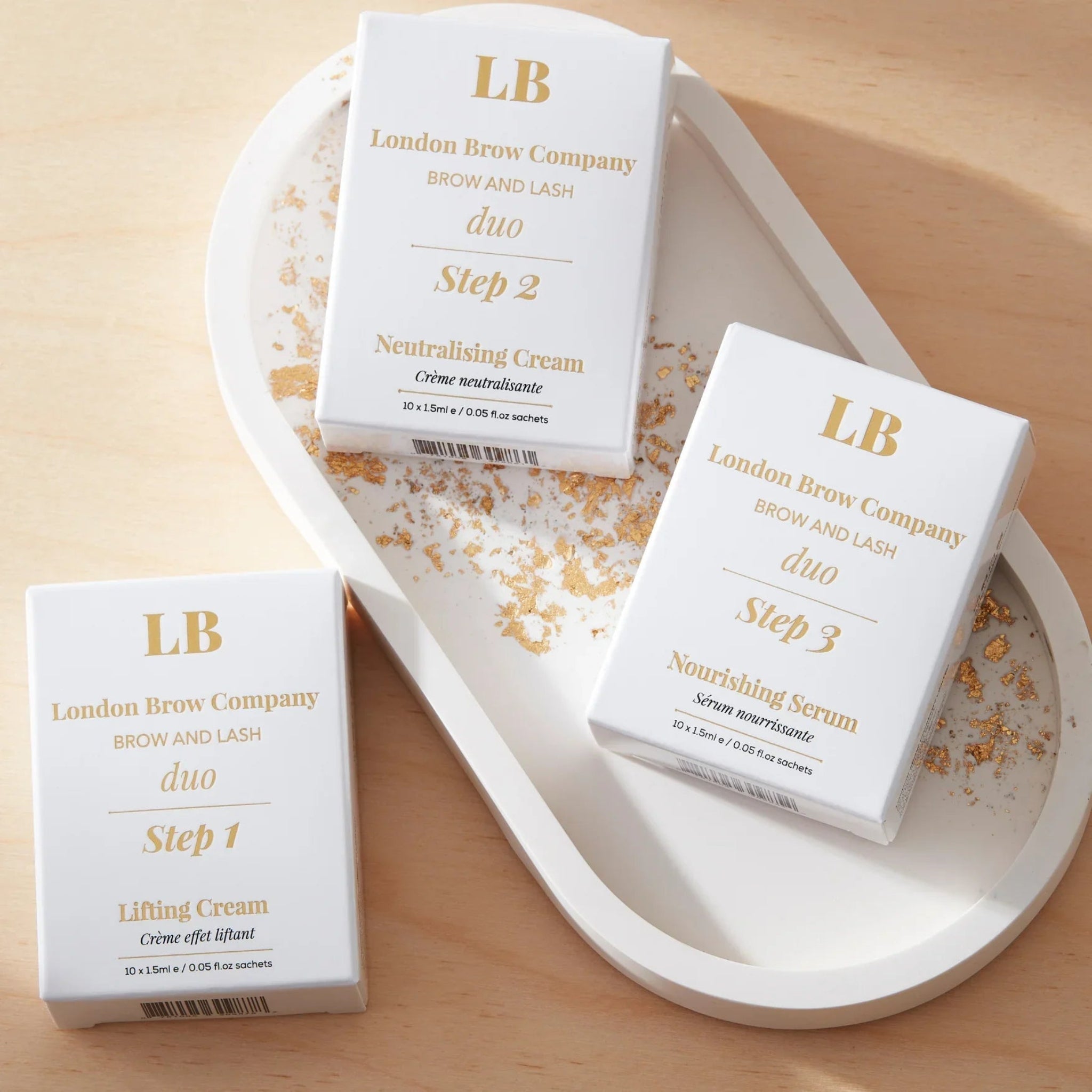 London Brow Company LB Brow and Lash Duo three-step sachet box set lifestyle product shot showing three white retail boxes labelled Step 1 Lifting Cream Step 2 Neutralising Cream and Step 3 Nourishing Serum each containing 10x1.5ml sachets with gold LB logo and London Brow Company Brow and Lash Duo branding arranged on a white oval ceramic tray with scattered gold leaf flakes on a warm wood surface from diagonal angle
