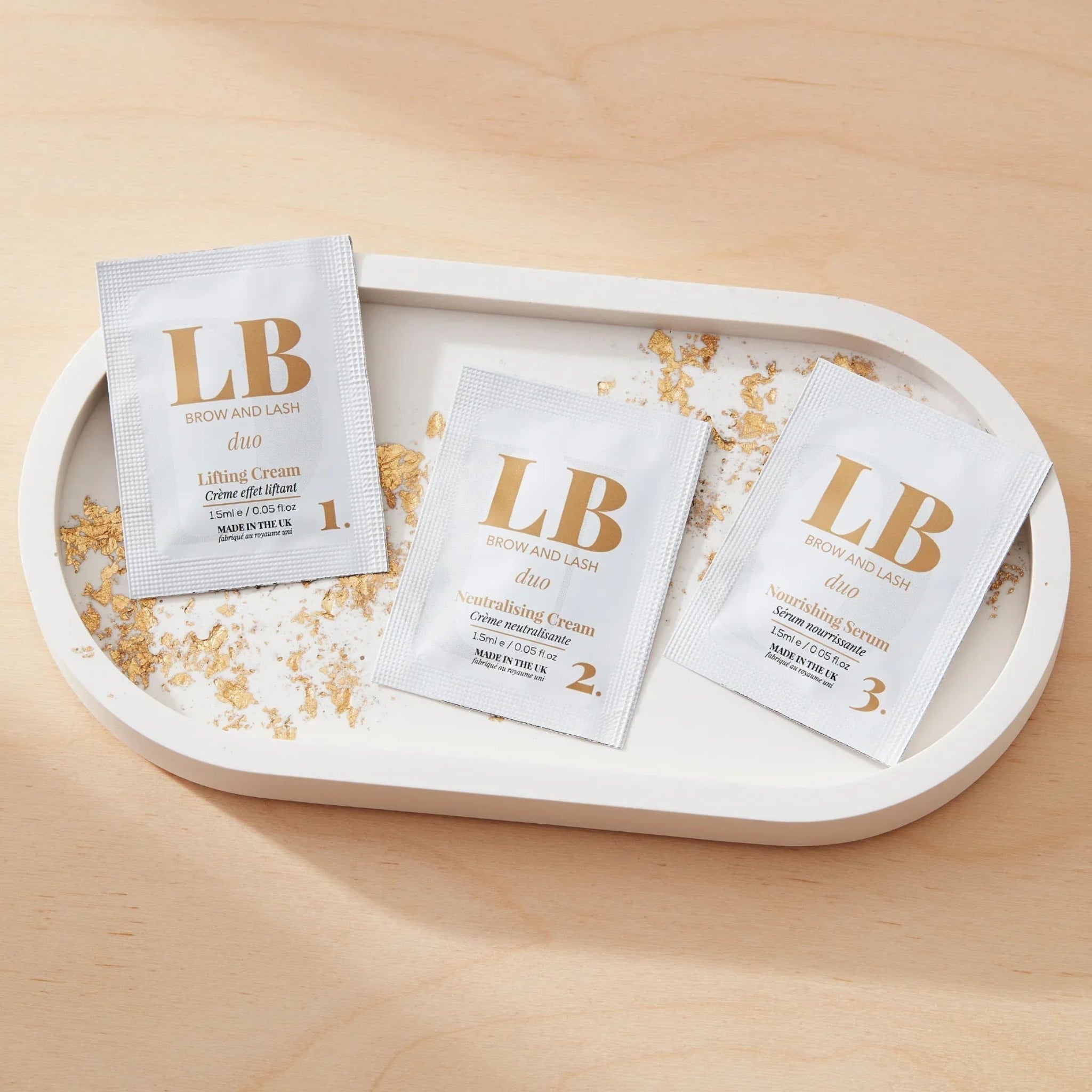 London Brow Company LB Brow and Lash Duo three-step sachet kit lifestyle flatlay showing three white foil sachets labelled Step 1 Lifting Cream Step 2 Neutralising Cream and Step 3 Nourishing Serum each 1.5ml Made in the UK arranged in a row on a white oval ceramic tray with scattered gold and white flakes on a warm wood surface from diagonal overhead angle