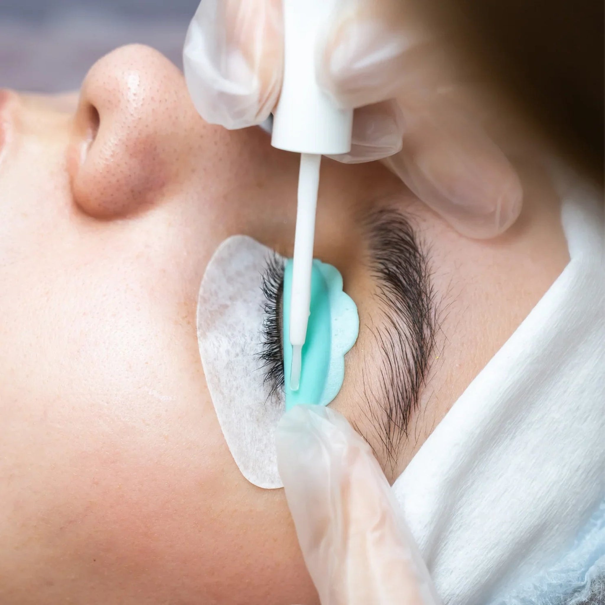 London Brow Company Speed-Lift Lash Lift and Tinting Course in-treatment close-up showing gloved therapist applying lash lift solution with a white micro applicator brush to natural lashes wrapped over a teal silicone lash shield on a fair-skinned female client with white under-eye pad from diagonal overhead angle