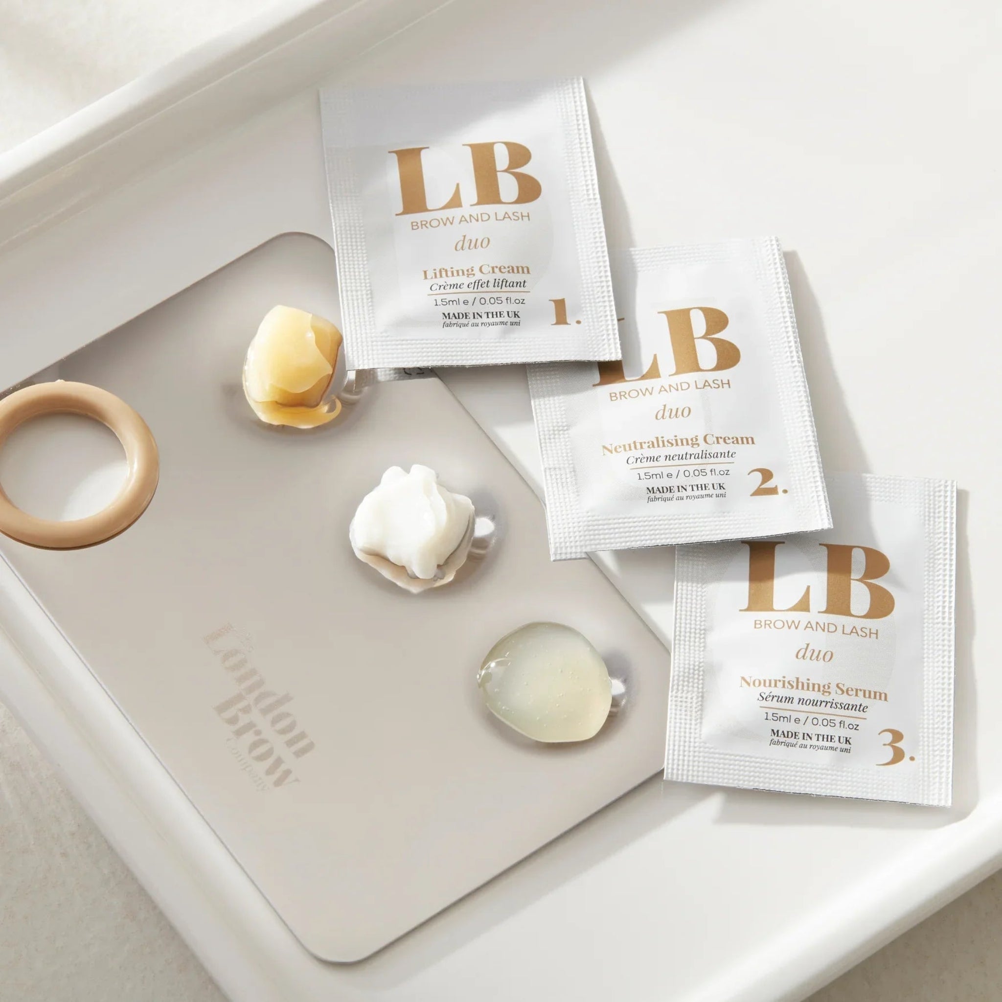 London Brow Company LB Brow and Lash Duo three-step sachet kit lifestyle flatlay showing three white foil sachets labelled Step 1 Lifting Cream Step 2 Neutralising Cream and Step 3 Nourishing Serum each 1.5ml Made in the UK alongside a beige glue ring two dollops of cream product and a clear serum drop on a grey London Brow Company silicone mat in a white ceramic tray from diagonal overhead angle
