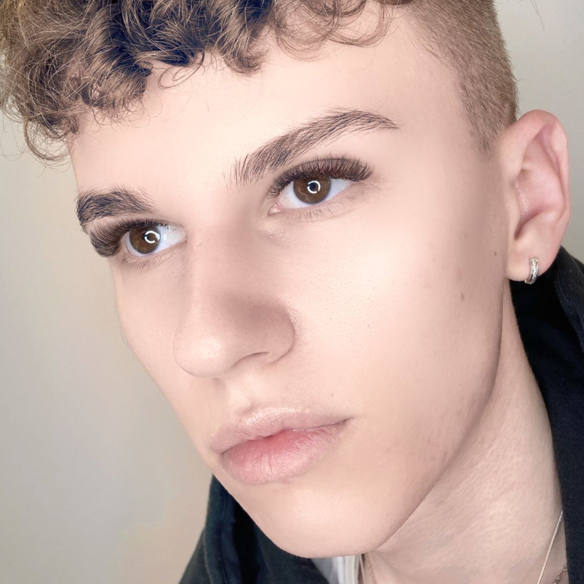 Eyelash Excellence Chocolate Brown YY Lashes model result shot showing young male model with curly blonde hair and brown eyes wearing natural chocolate brown YY lash extensions and defined brows from three-quarter angle with silver hoop earring visible