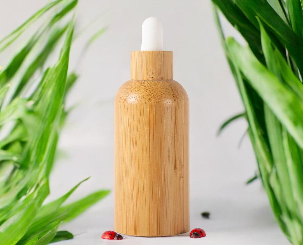 A 60ml bamboo eyelash rinse bottle with a clear pipette, positioned on a white surface with green plant leaves and a few red berries nearby.