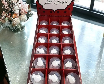 A display box containing 18 bottles of lash shampoo, with the product label 'Lash Shampoo Bubble Gum' on the top. The box is presented on a table with a floral decoration in the background.