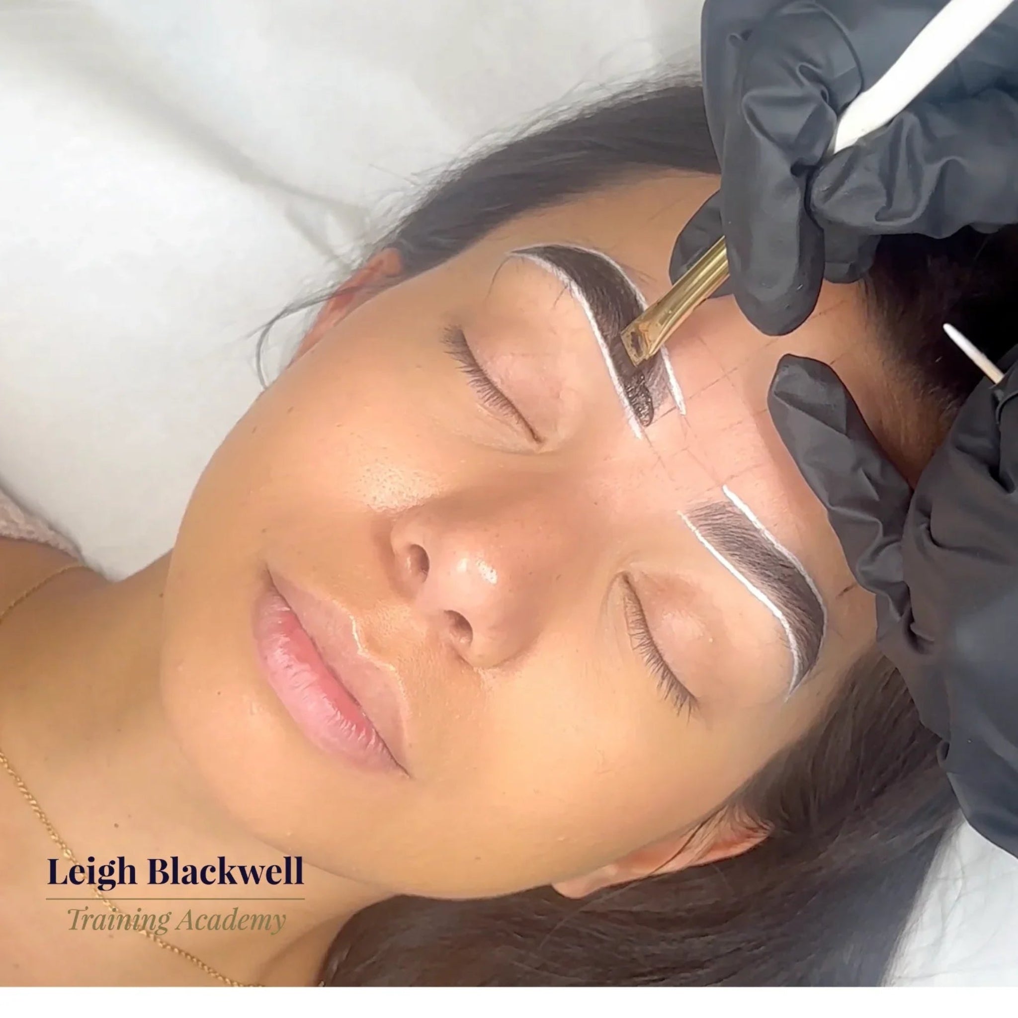 London Brow Company Liquid Stain Eyebrow Tinting Course in-treatment shot showing gloved therapist in black latex gloves applying dark liquid brow stain to a mapped and outlined brow on a reclined olive-skinned female client using a gold-handled flat brush from diagonal overhead angle on white treatment bed