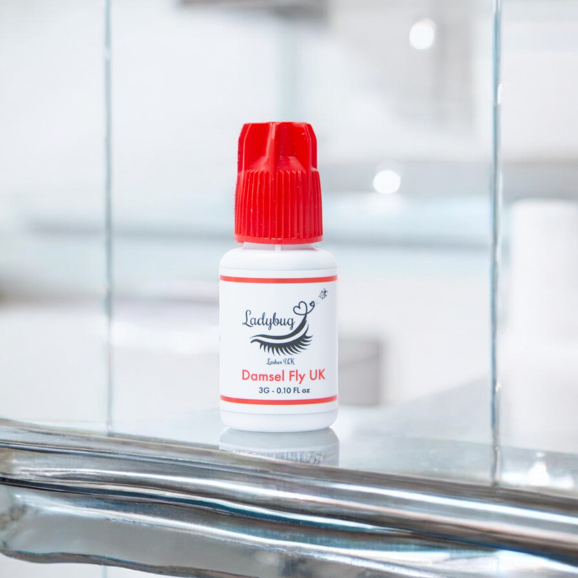 A bottle of Damselfly UK adhesive/glue with a red cap on a reflective surface, labelled for professional use by Ladybug Lashes.