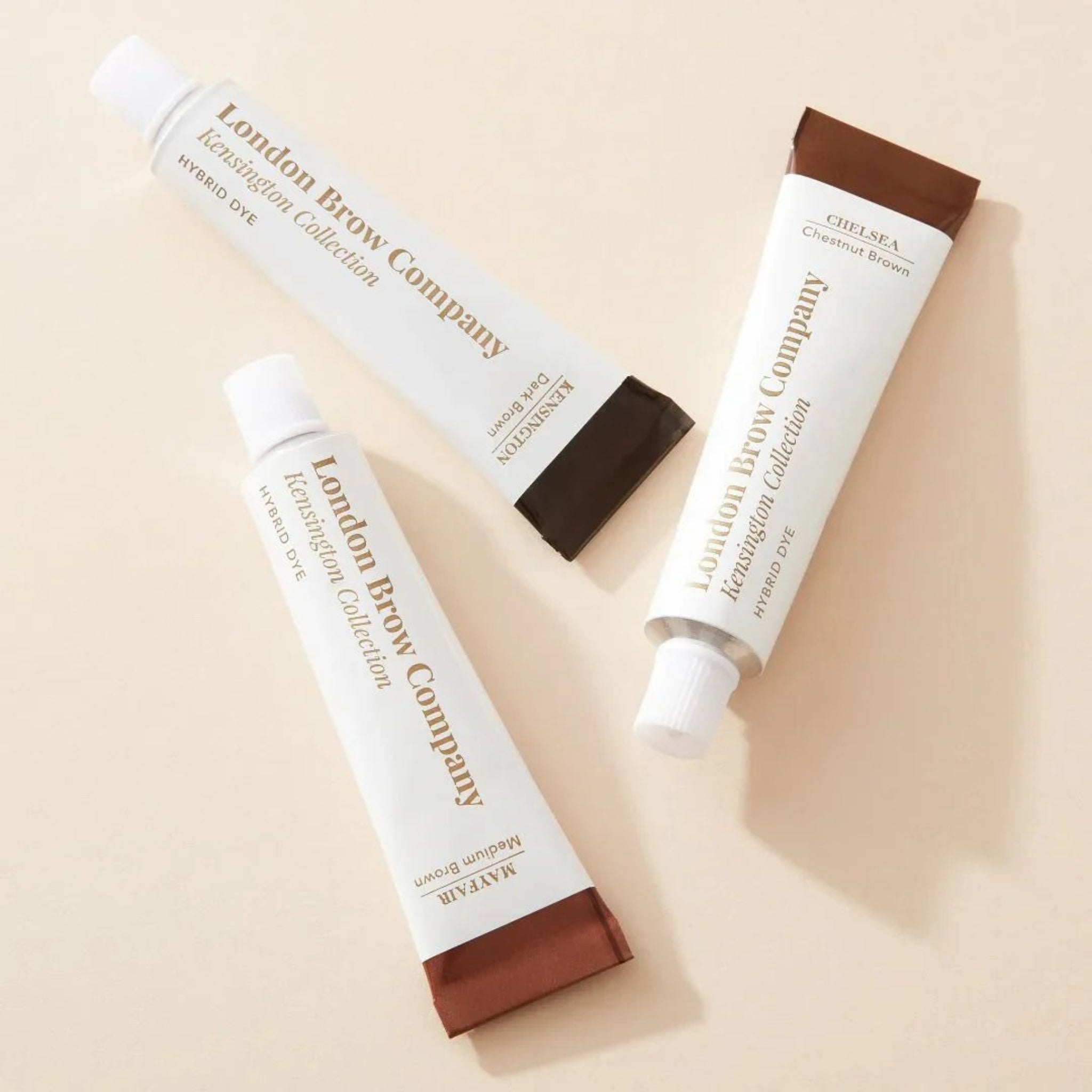 London Brow Company Kensington Collection Hybrid Dye tubes close-up flatlay showing three white squeeze tubes labelled Kensington Dark Brown Mayfair Medium Brown and Chelsea Chestnut Brown with colour-coded caps in dark brown and chestnut brown from diagonal overhead angle on warm cream background