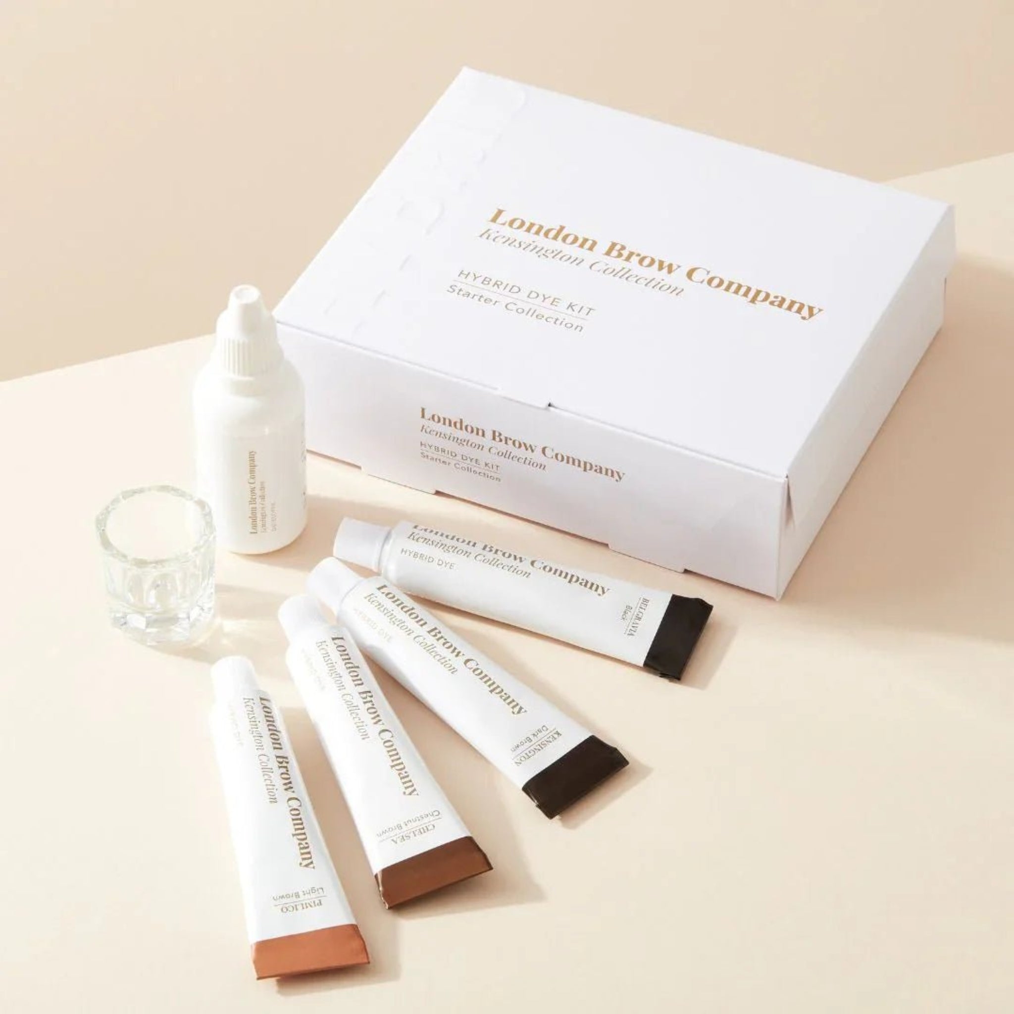 London Brow Company Kensington Collection Hybrid Dye Kit Starter Collection lifestyle product shot showing white branded box open with four hybrid dye tubes in brown shades developer bottle and clear mixing cup arranged on a warm cream surface from diagonal angle