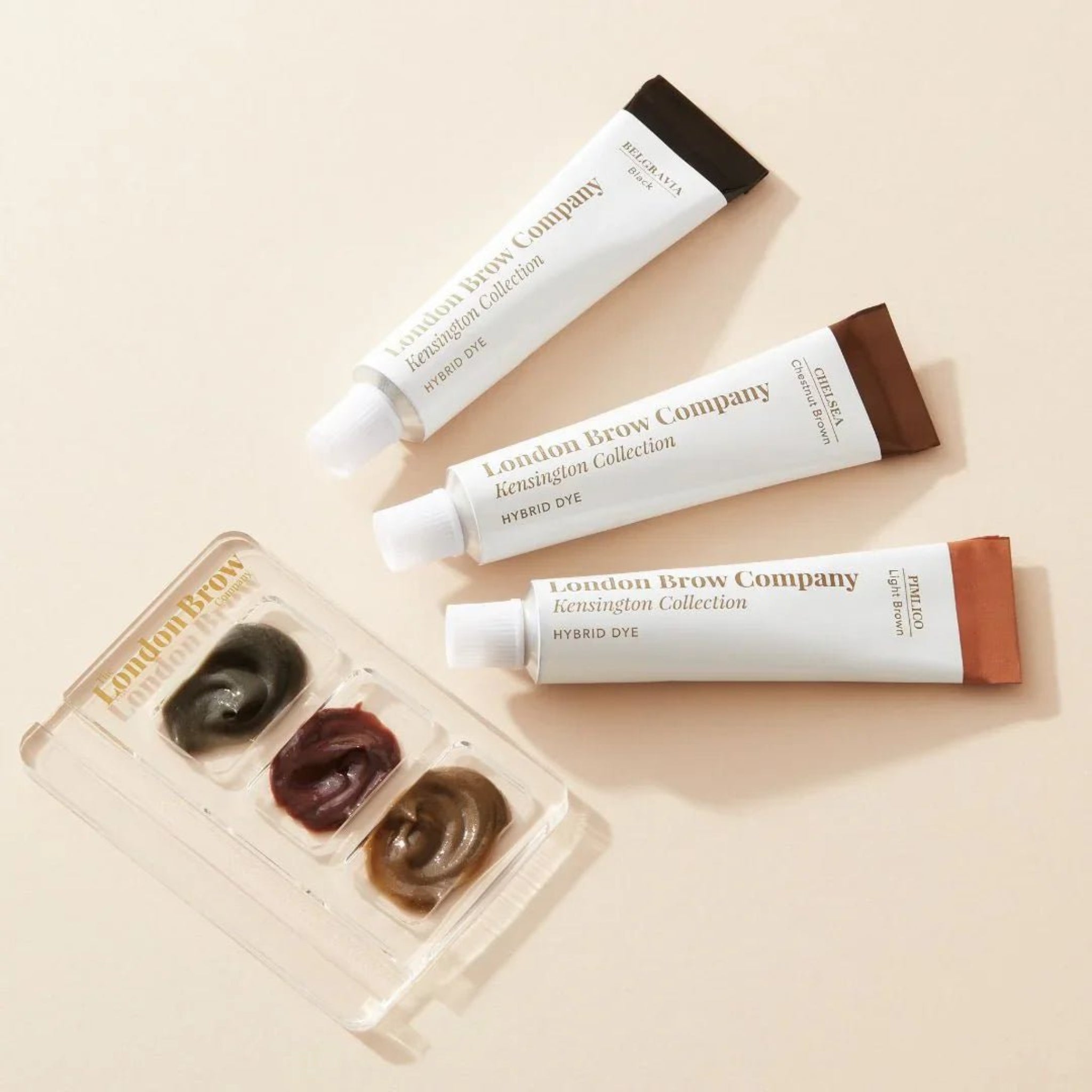 London Brow Company Kensington Collection Hybrid Dye Starter Kit lifestyle flatlay showing three white Hybrid Dye tubes in Belgravia Black Chelsea Chestnut Brown and Pimlico Light Brown shades fanned out alongside a London Brow Company clear acrylic mixing palette with three mixed dye colour swatches from diagonal overhead angle on warm cream background