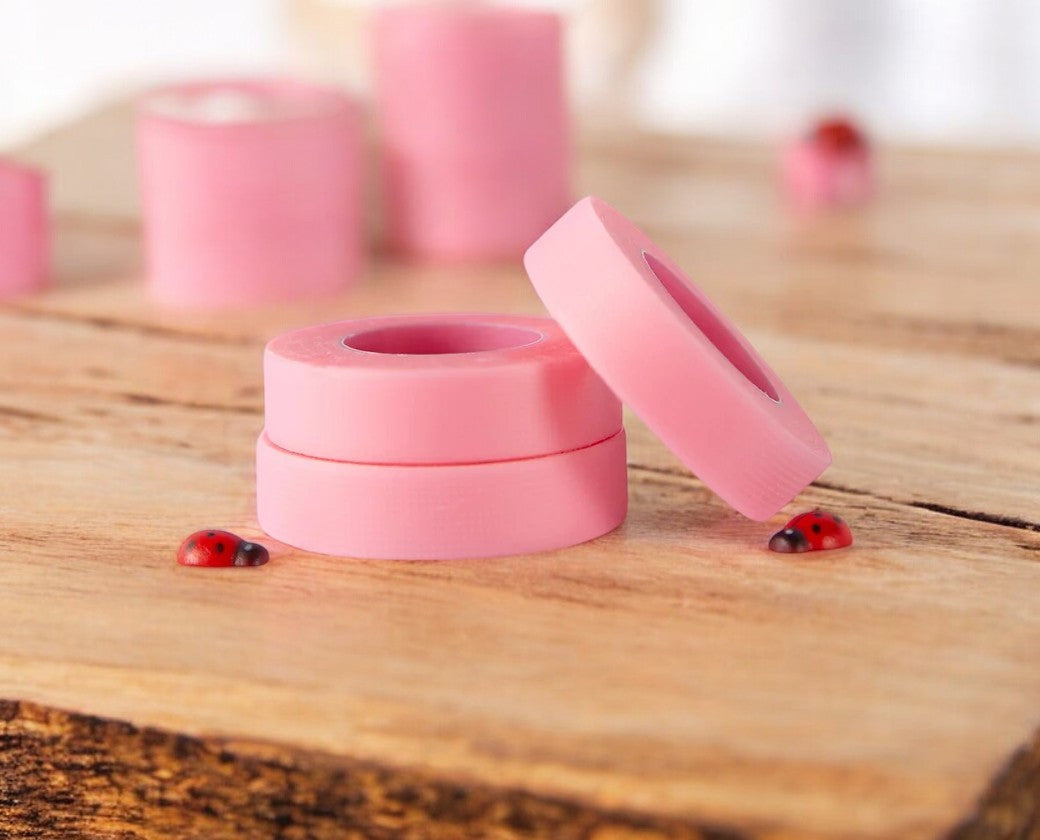 A roll of pink silicone tape for eyelash extension applications, placed on a wooden surface with a few eyelashes and a ladybug in the background.