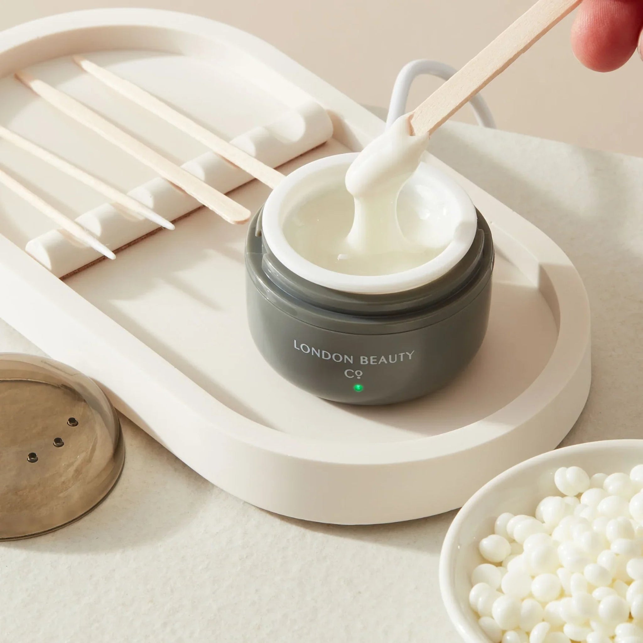 London Beauty Co Mini Portable Wax Heater in-use lifestyle shot showing dark grey USB-powered mini wax heater with green indicator light on white oval tray with a hand dipping a wooden spatula into melted white wax alongside additional wooden spatulas and a bowl of white wax pellets from diagonal overhead angle on cream background