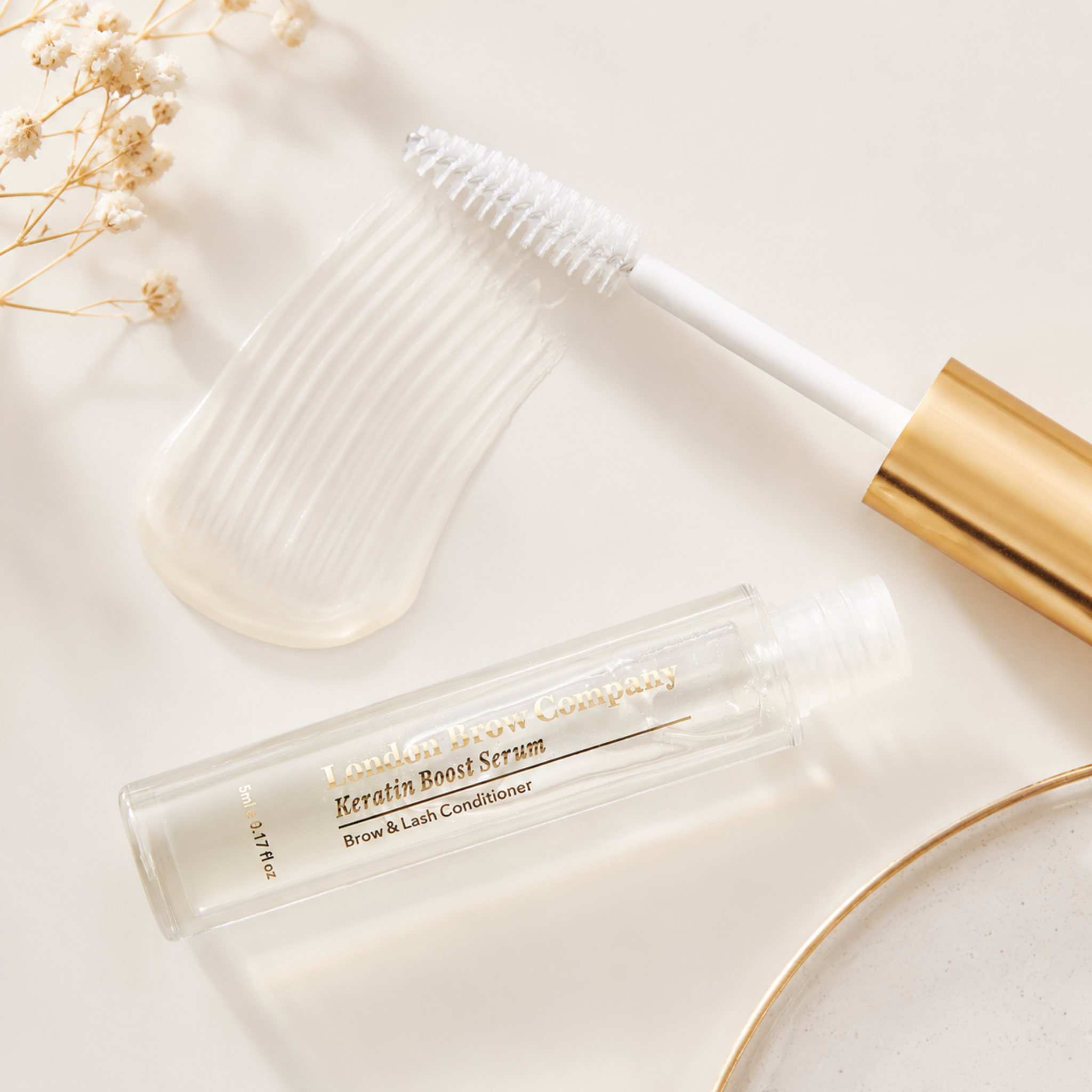 The London Brow Company Keratin Brow & Lash Boost Serum flatlay product shot showing clear serum tube with gold cap and open spoolie brush from overhead angle on cream background with dried flowers and gold dish