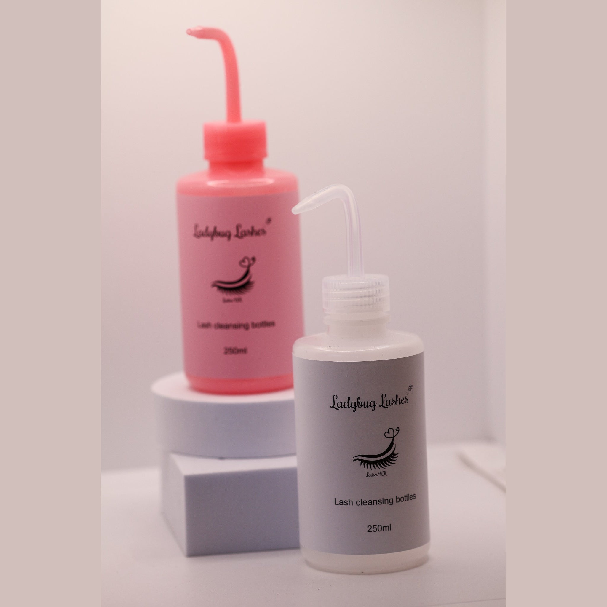 Pink and clear Ladybug Lashes 250ml lash cleansing rinse bottles displayed together on white geometric risers against a white background