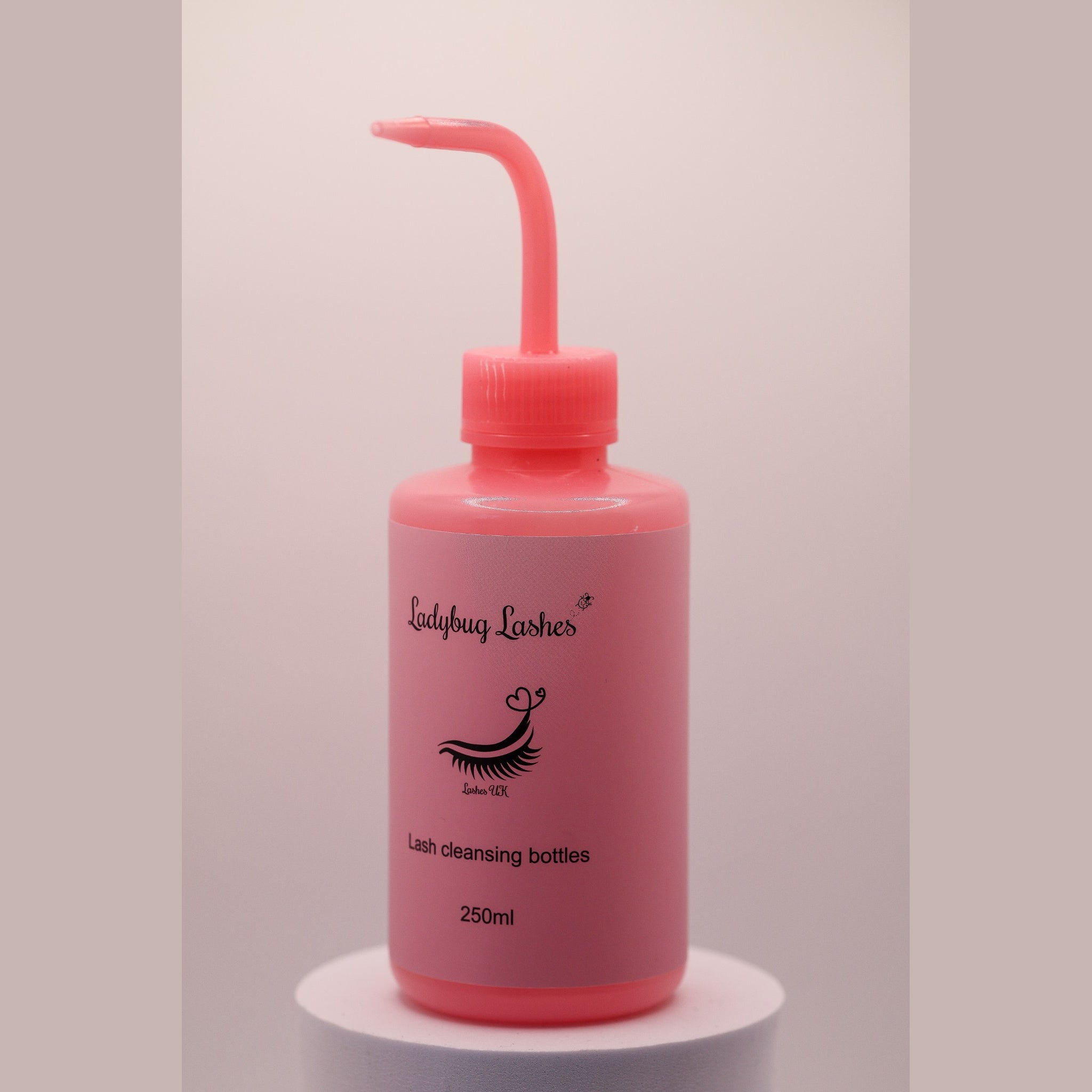 Pink Ladybug Lashes 250ml lash cleansing rinse bottle with angled nozzle tip, shown upright on a white plinth against a soft pink background