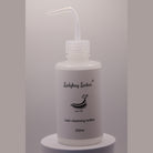 Clear Ladybug Lashes 250ml lash cleansing rinse bottle with angled nozzle tip, shown upright on a white plinth against a grey background