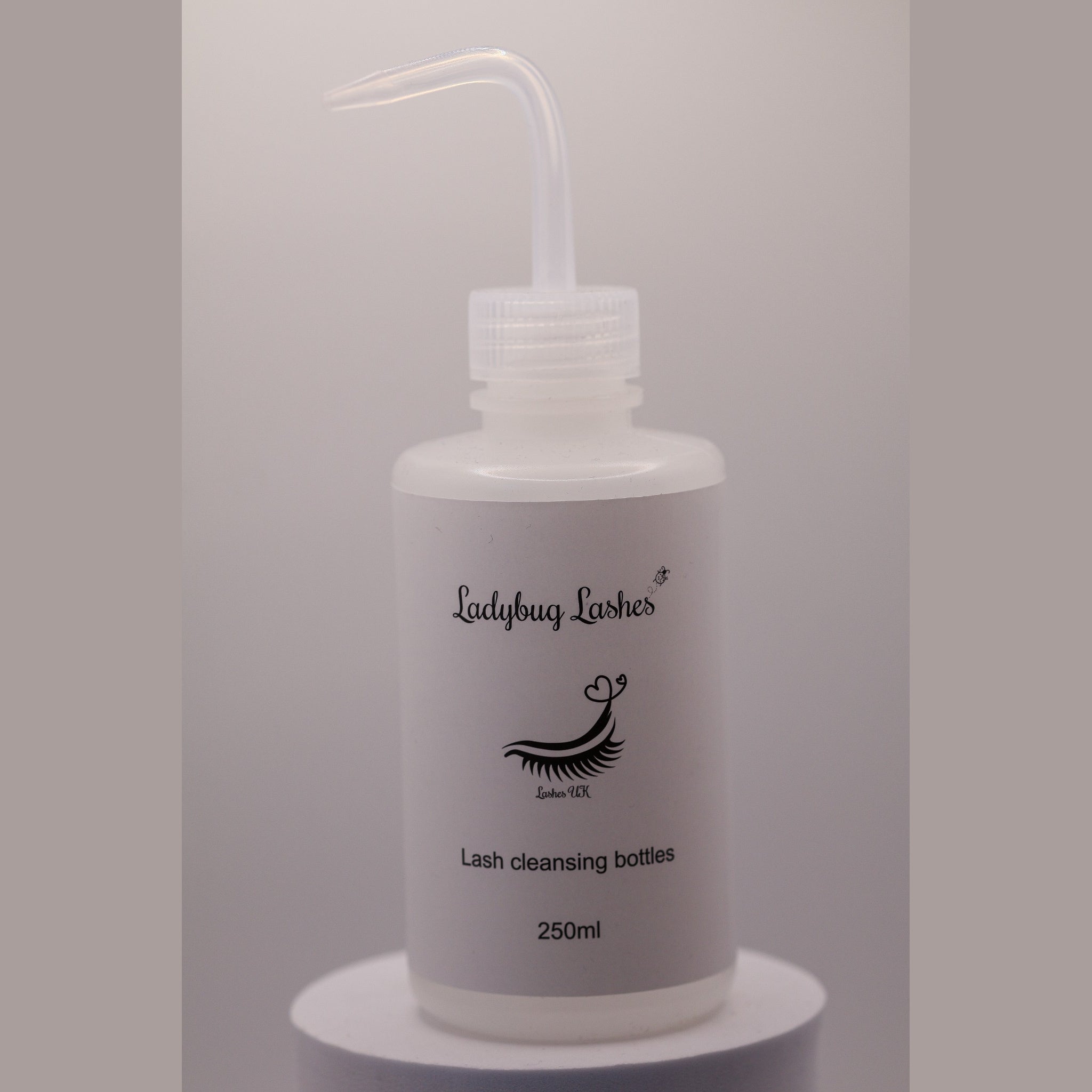 Clear Ladybug Lashes 250ml lash cleansing rinse bottle with angled nozzle tip, shown upright on a white plinth against a grey background