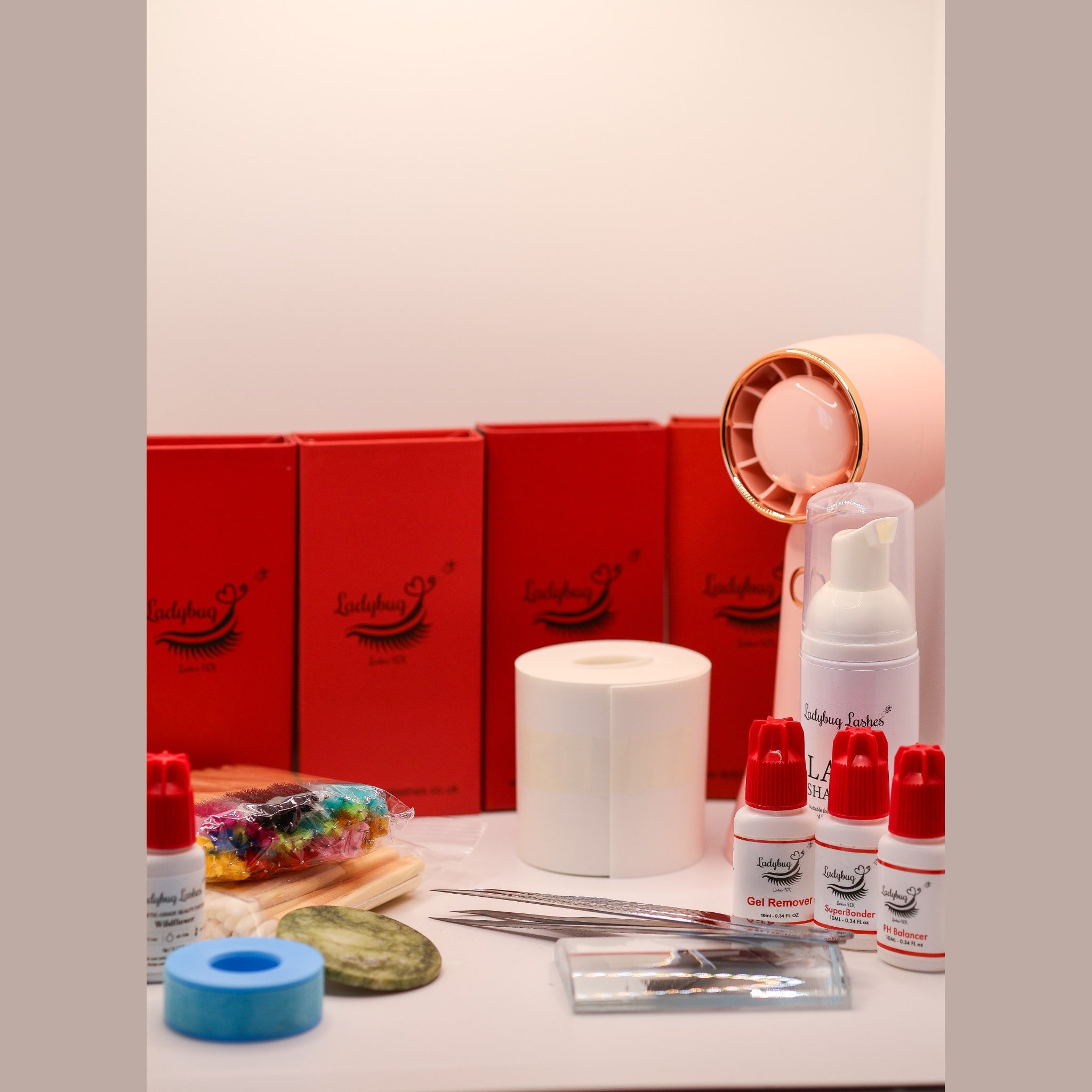Ladybug Lashes Complete Gold Lash Starter Kit flatlay showing four red Ladybug Lashes lash tray boxes, white foam lash shampoo pump bottle, three white and red Ladybug Lashes bottles labelled Gel Remover, SuperBonder and pH Balancer, adhesive bottle, roll of white under-eye pads, blue gel tape roll, jade stone, glass lash palette, isolation tweezers, coloured mascara spoolies and a pink rechargeable USB mini fan on a white surface