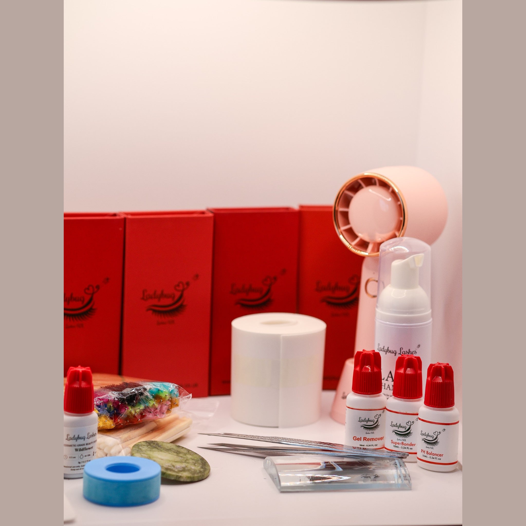 Ladybug Lashes Complete Gold Lash Starter Kit contents displayed on a white surface showing four red Ladybug Lashes lash tray boxes, white foam lash shampoo pump bottle, three white and red Ladybug Lashes bottles labelled Gel Remover, SuperBonder and pH Balancer, adhesive bottle, roll of white under-eye pads, blue gel tape roll, jade stone, glass lash palette, isolation tweezers, coloured mascara spoolies on a jade stone and a pink rechargeable USB mini fan