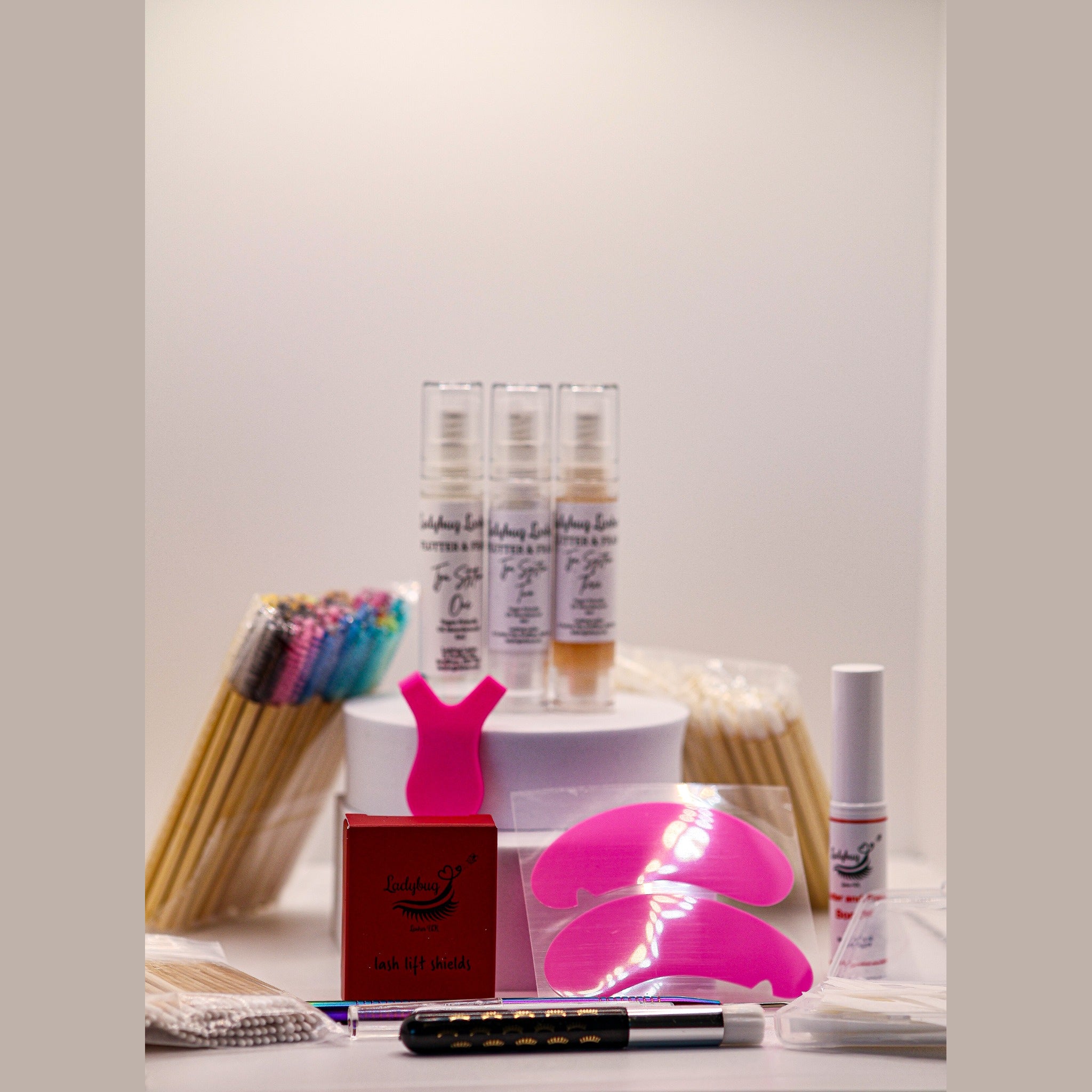 Ladybug Lashes Flutter and Frame Airless Pump Lash Lift Kit contents displayed on a white surface showing three Ladybug Lashes Flutter and Frame Step One Two Three airless pump bottles on a white plinth, pink Y-shaped silicone lifting tool, Ladybug Lashes lash lift glue balm pot, red Ladybug Lashes branded box, coloured mascara spoolies, wooden spatulas, lash lifting rod, fine detail brushes and a clear plastic storage case