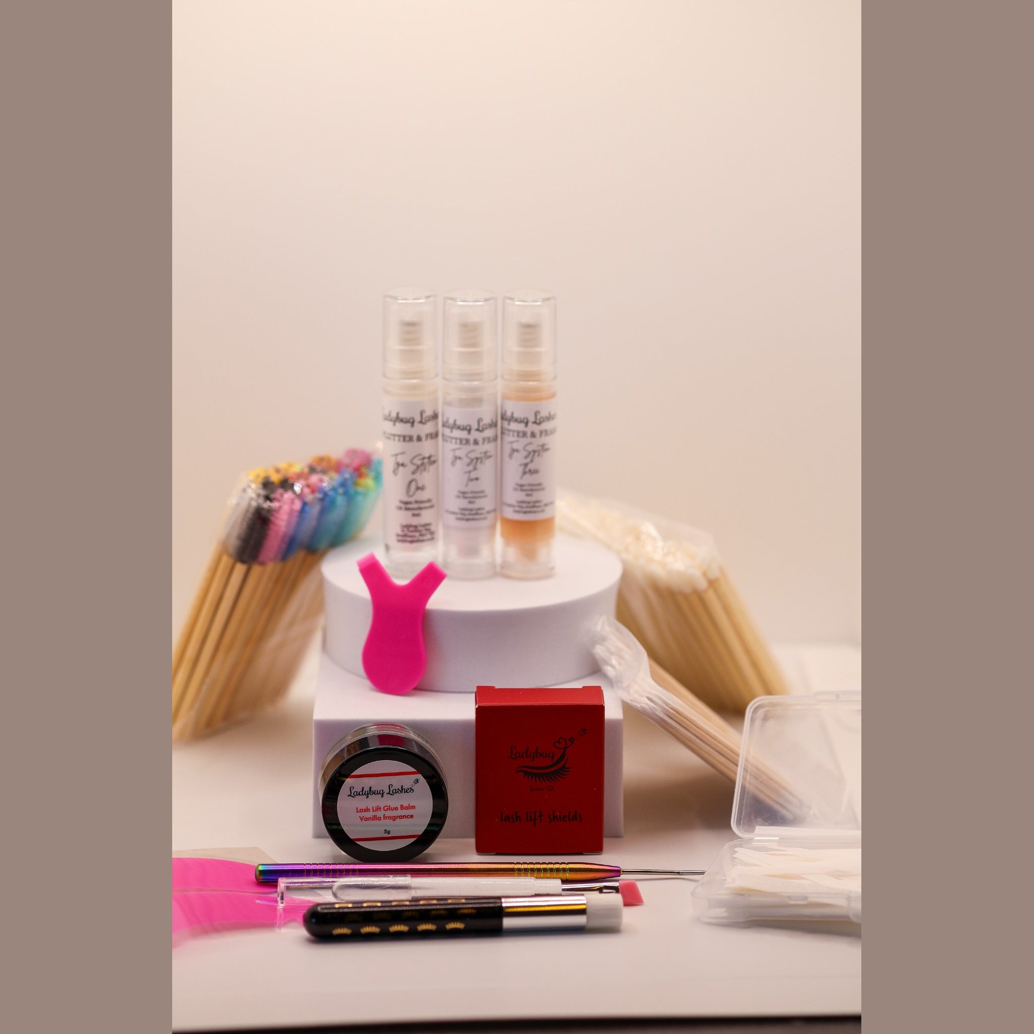 Ladybug Lashes Flutter and Frame Airless Pump Lash Lift Kit flatlay showing three Ladybug Lashes Flutter and Frame Step One Two Three airless pump bottles on a white plinth, pink Y-shaped silicone lifting tool, red Ladybug Lashes branded box, pink silicone under-eye patches, white lash shampoo bottle, coloured mascara spoolies, wooden spatulas, lash lifting rod, fine detail brushes and cotton buds on a white surface