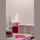 Ladybug Lashes Flutter and Frame Lash Lift Sachet Kit contents displayed on a white surface showing three Ladybug Lashes Flutter and Frame Tga System One Two Three sachet boxes, Ladybug Lashes lash lift glue balm pot, red Ladybug Lashes branded box, pink Y-shaped silicone lifting tool, coloured mascara spoolies in a bag, paper compensators, pink silicone under-eye patches, fine detail brushes and a clear plastic storage case
