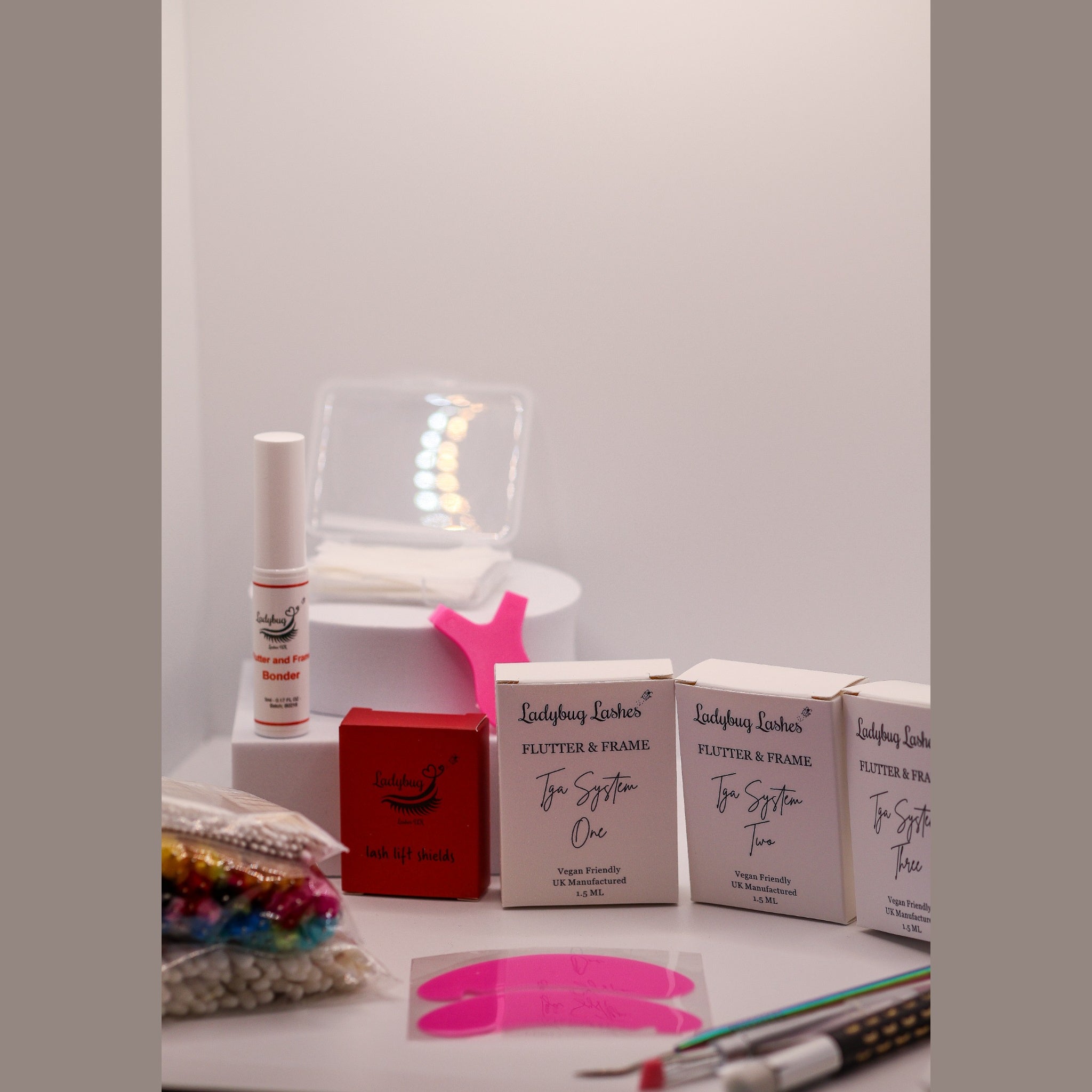 Ladybug Lashes Flutter and Frame Lash Lift Sachet Kit contents displayed on a white surface showing three Ladybug Lashes Flutter and Frame Tga System One Two Three sachet boxes, Ladybug Lashes lash lift glue balm pot, red Ladybug Lashes branded box, pink Y-shaped silicone lifting tool, coloured mascara spoolies in a bag, paper compensators, pink silicone under-eye patches, fine detail brushes and a clear plastic storage case