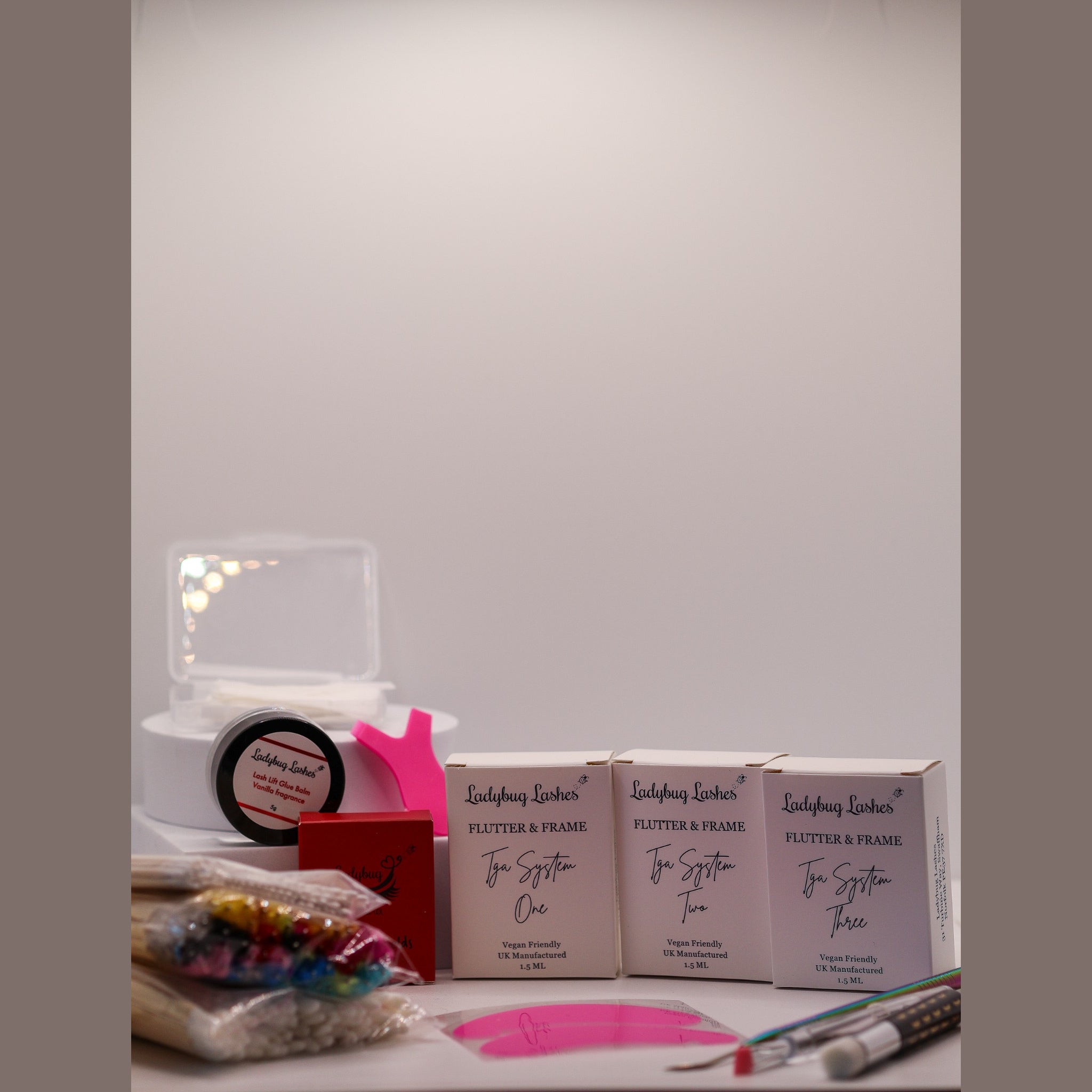 Ladybug Lashes Flutter and Frame Lash Lift Sachet Kit flatlay showing three Ladybug Lashes Flutter and Frame Tga System One Two Three sachet boxes, white lash adhesive bottle, red Ladybug Lashes branded box, pink Y-shaped silicone lifting tool, pink silicone under-eye patches, coloured mascara spoolies in a bag, paper compensators, fine detail brushes and a white plinth on a white surface
